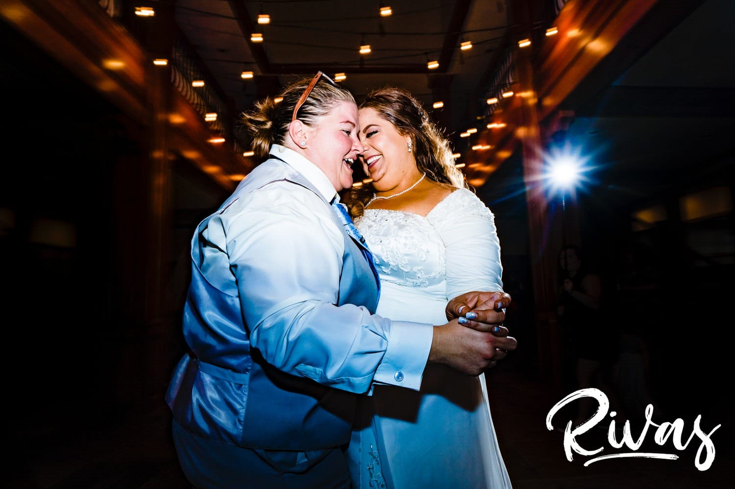 A colorful, wide picture of two brides holding hands as they dance together underneath a canopy of twinkly lights during their wedding reception at Fire Stables. 