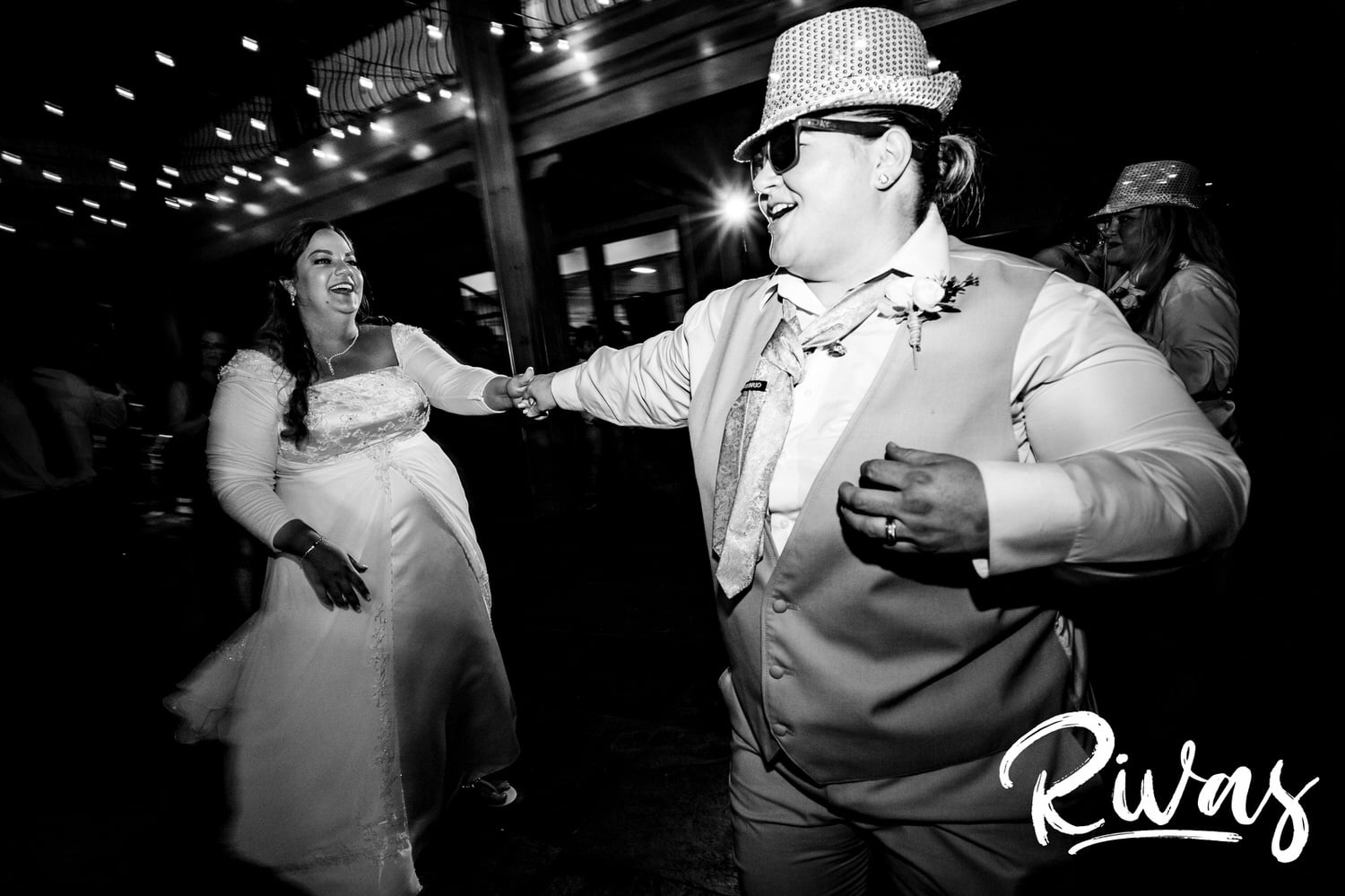 A candid black and white picture of a bride in a sequined fedora spinning her bride across the dance floor during their wedding reception at Fire Stables. 
