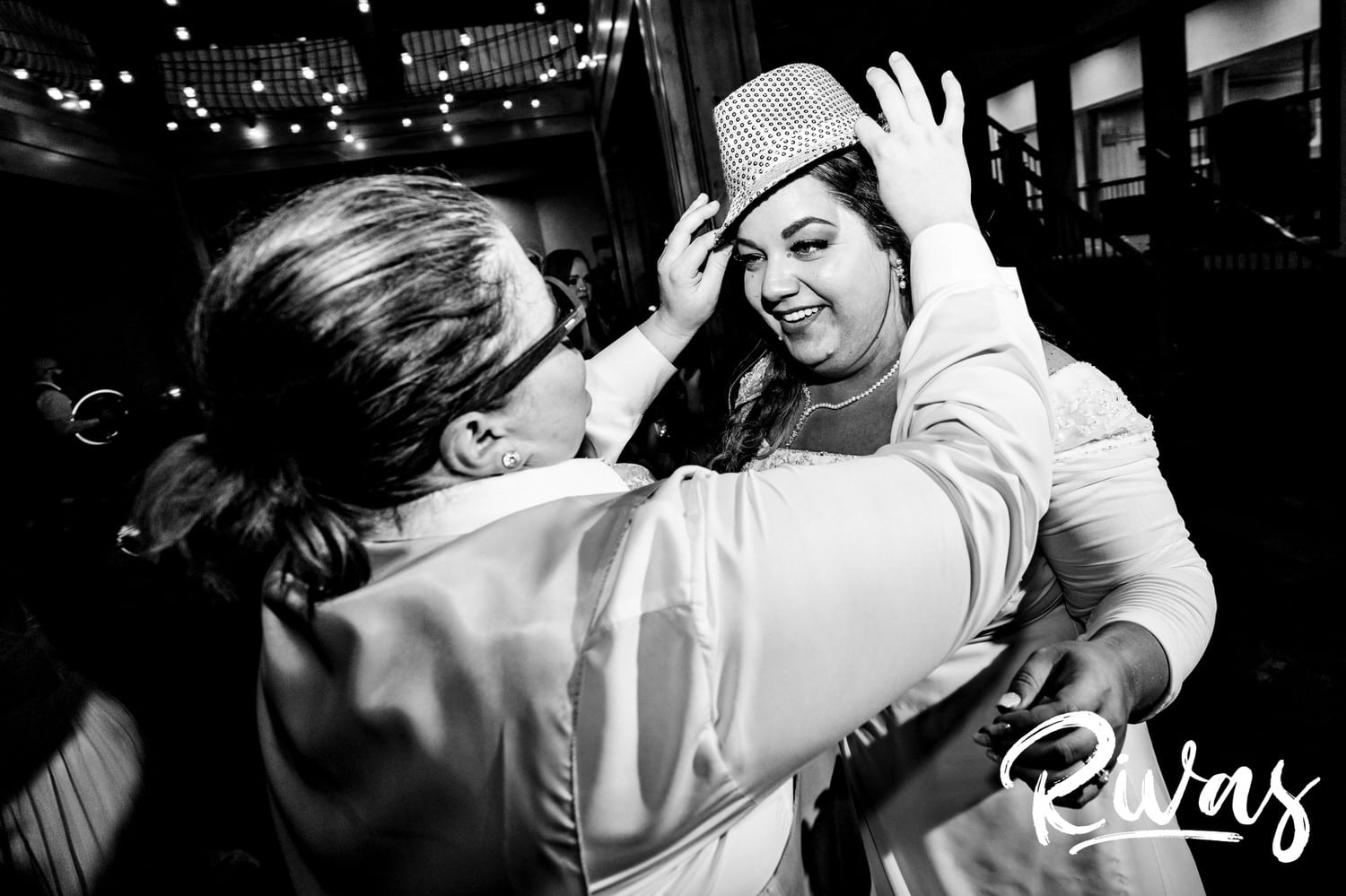 A candid black and white picture of a bride adjusting a sequined fedora on her wife's head during their Fire Stables wedding reception. 