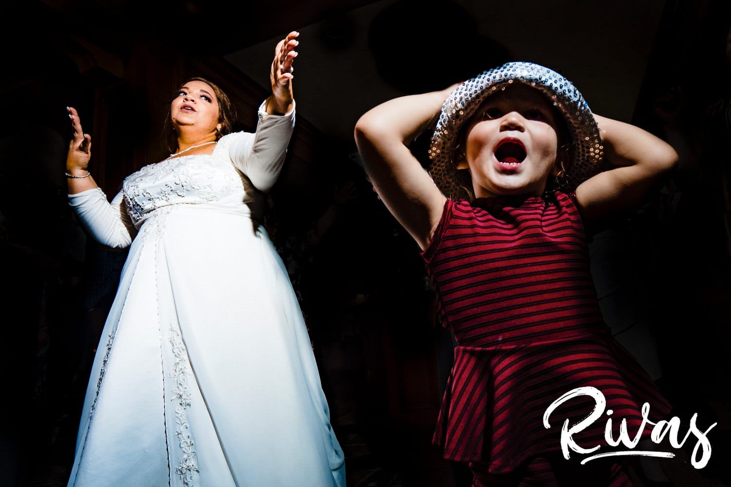 A candid picture of a bride dancing enthusiastically as a little girl in a sequined hat sings excitedly during a wedding reception at Fire STables. 