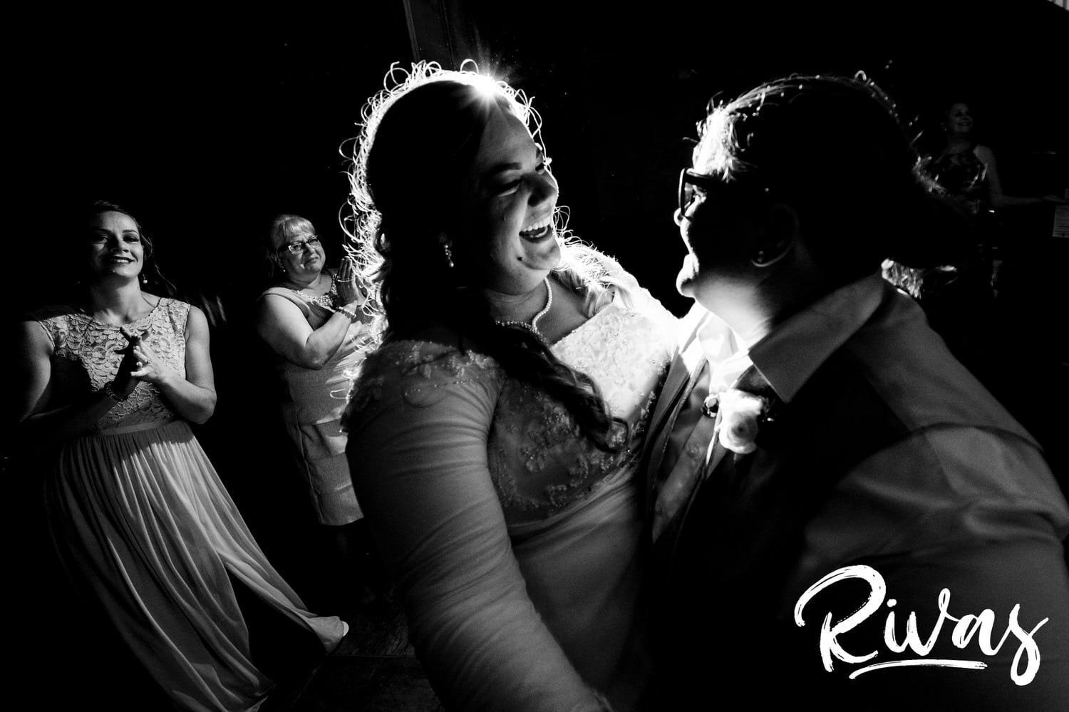A candid black and white picture of two brides dancing together as their friends and family look on. 