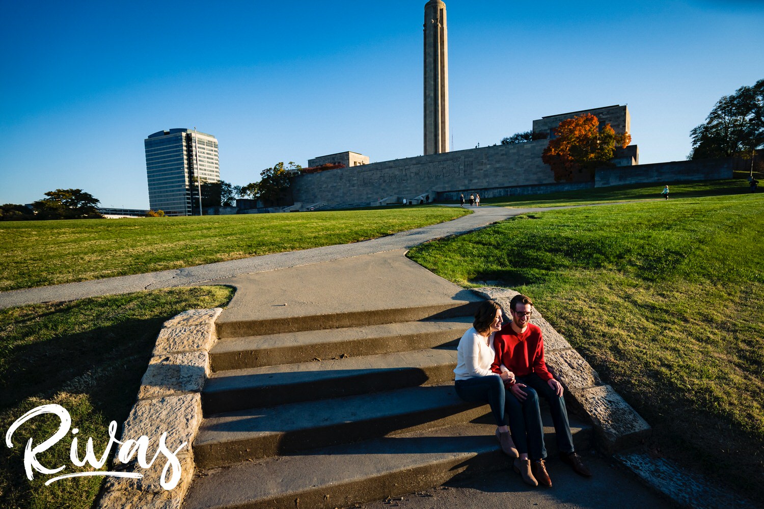 I wide portrait of an engaged couple sitting on a set of stairs on the lawn in front of Liberty Memorial during their fall engagement session in Kansas City. 