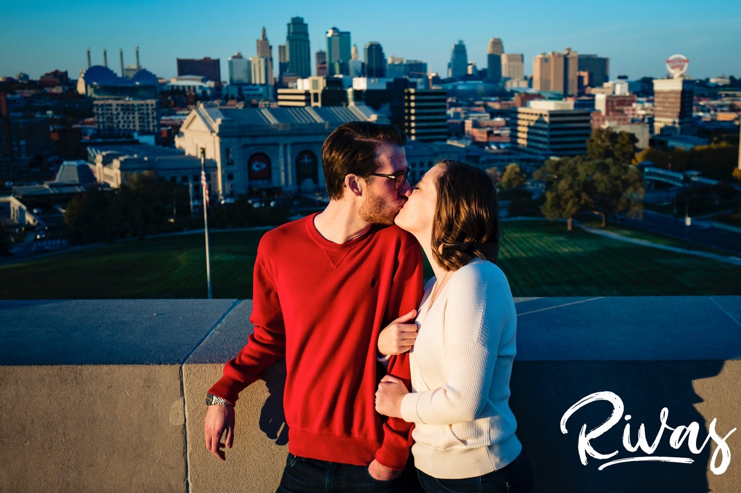 A candid, close-up picture of an engaged couple sharing a kiss as they stand in front of the Kansas City skyline at Liberty Memorial during their engagement session. 