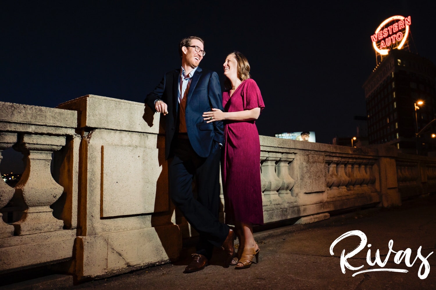 A candid portrait of an engaged couple leaning up against a stone colonnade with the Western Auto sign lit up behind them during their Kansas City engagement session. 