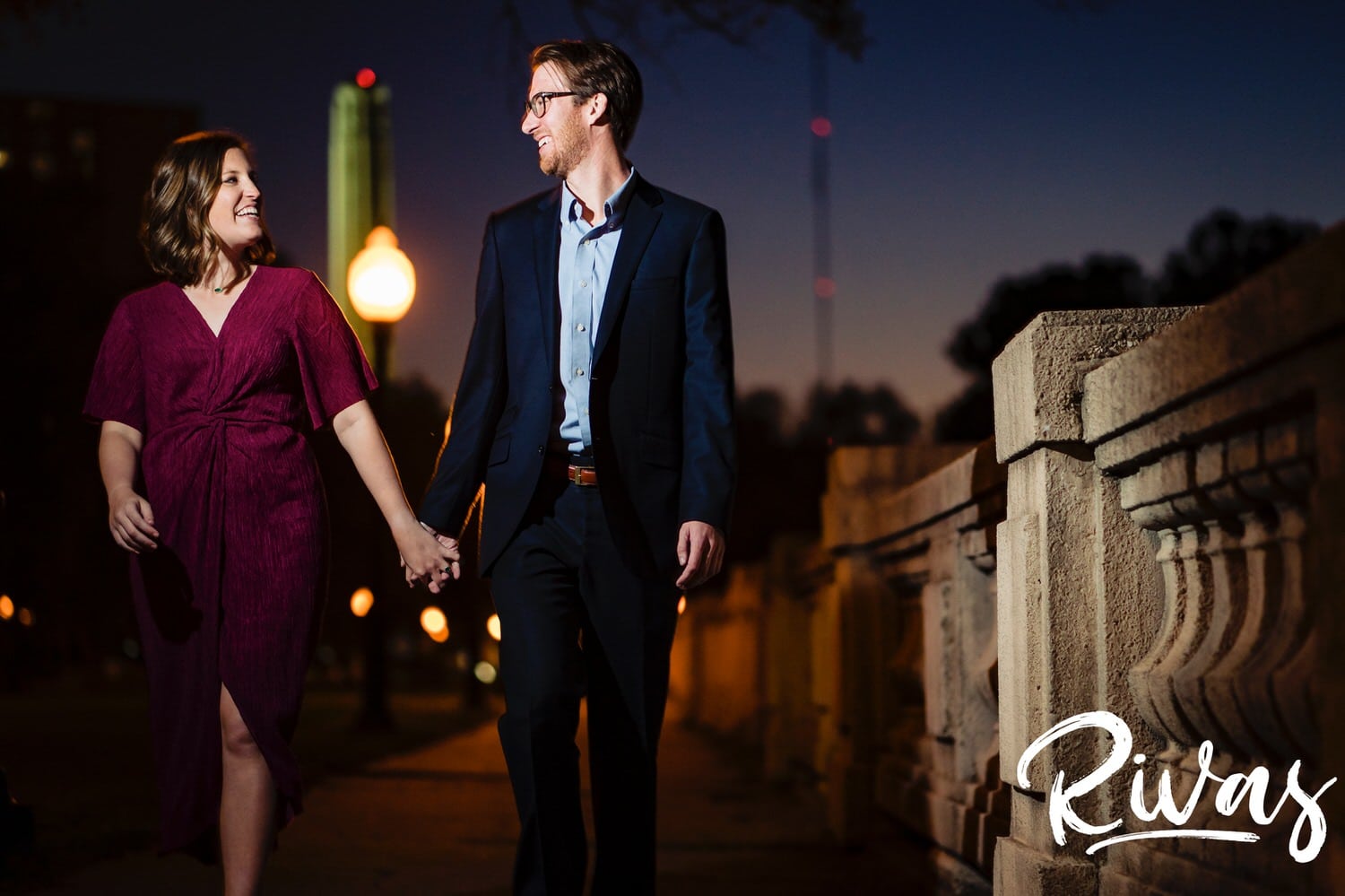 A picture of an engaged couple holding hands walking on a park just after dusk with Liberty Memorial visible in the background during their Kansas City Engagement Session. 