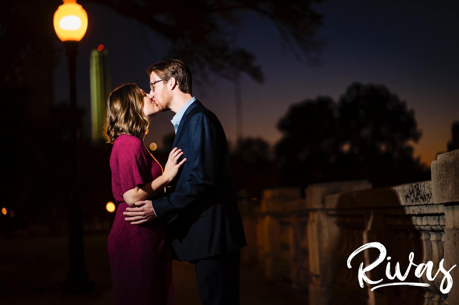 A candid picture of an engaged couple leaning in to share a kiss just after sun set with Liberty Memorial visible behind them during their engagement session in Kansas City. 