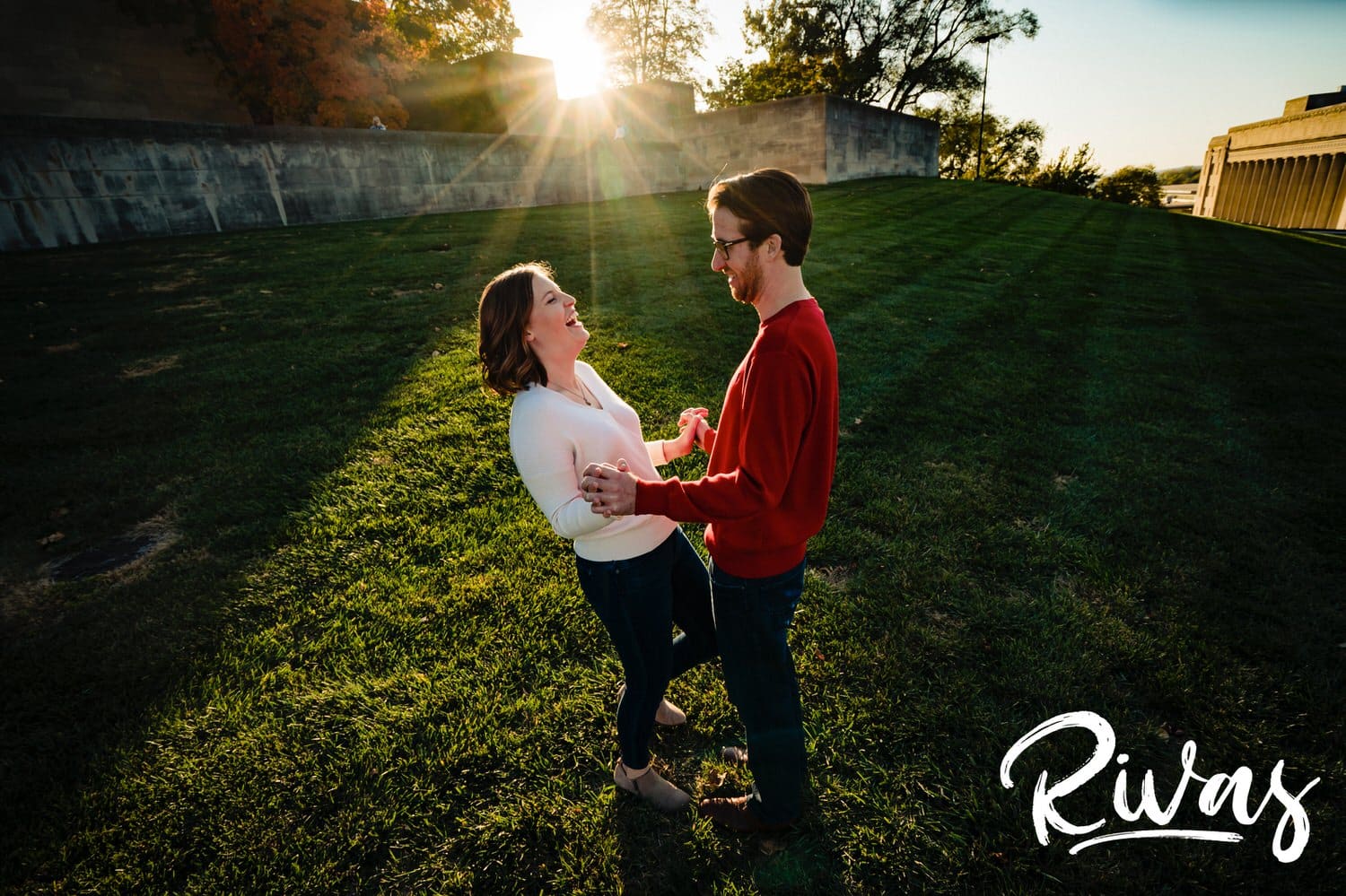 A candid picture of an engaged couple holding hands and laughing together standing on a green lawn with colorful autumn leaves in the background during their engagement session at Liberty Memorial in Kansas City. 