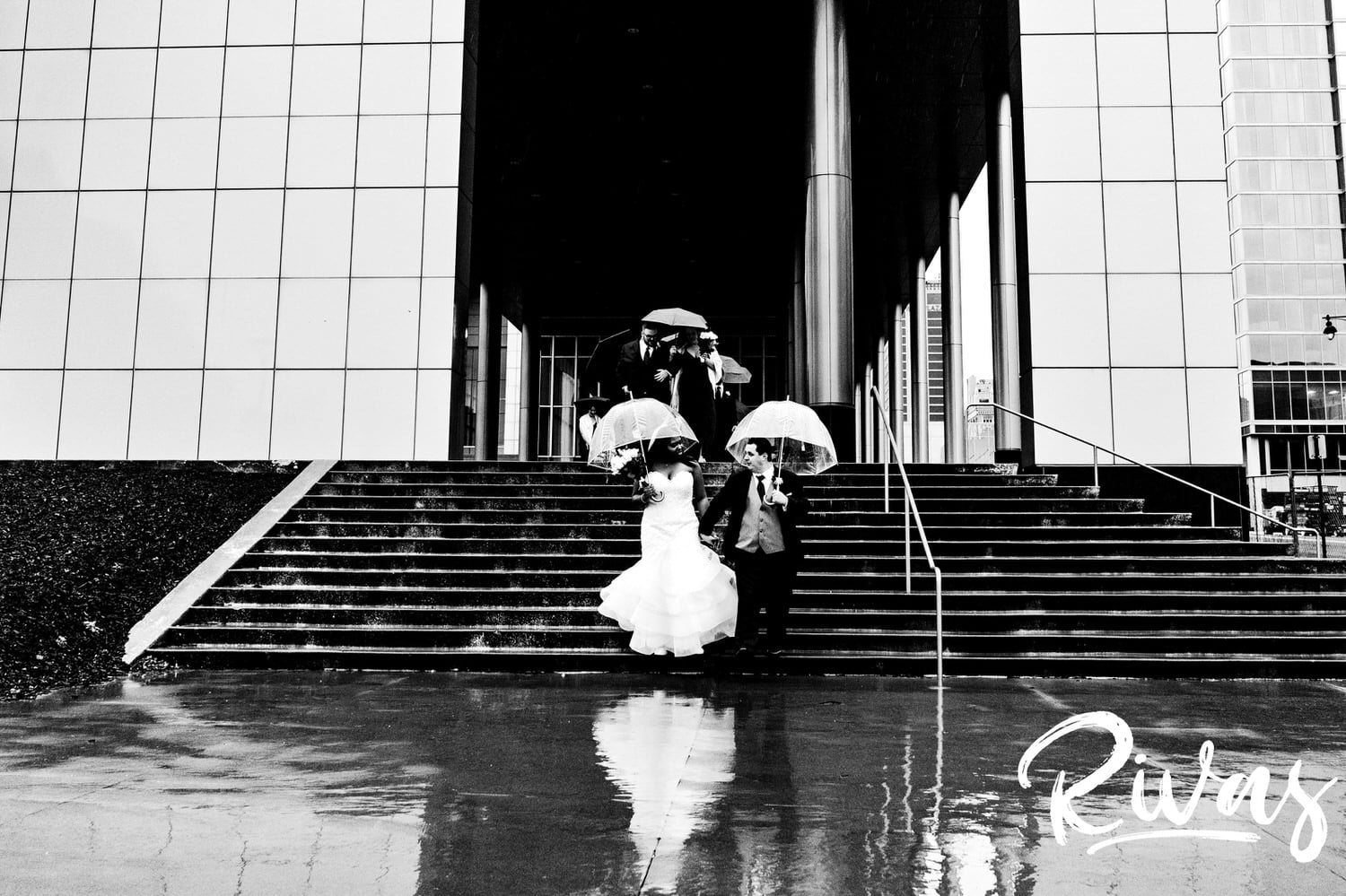 A candid black and white picture of a bride and groom with clear umbrellas running down a set of stairs at The Kansas City Convention Center in the rain on their December wedding day in Kansas City.