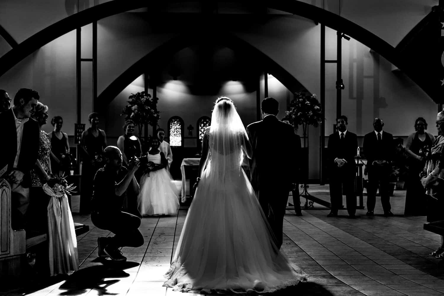 A candid, black and white picture of a bride and her father walking down the aisle with a photographer on the side taking a picture of them during a summer wedding ceremony in Kansas City.