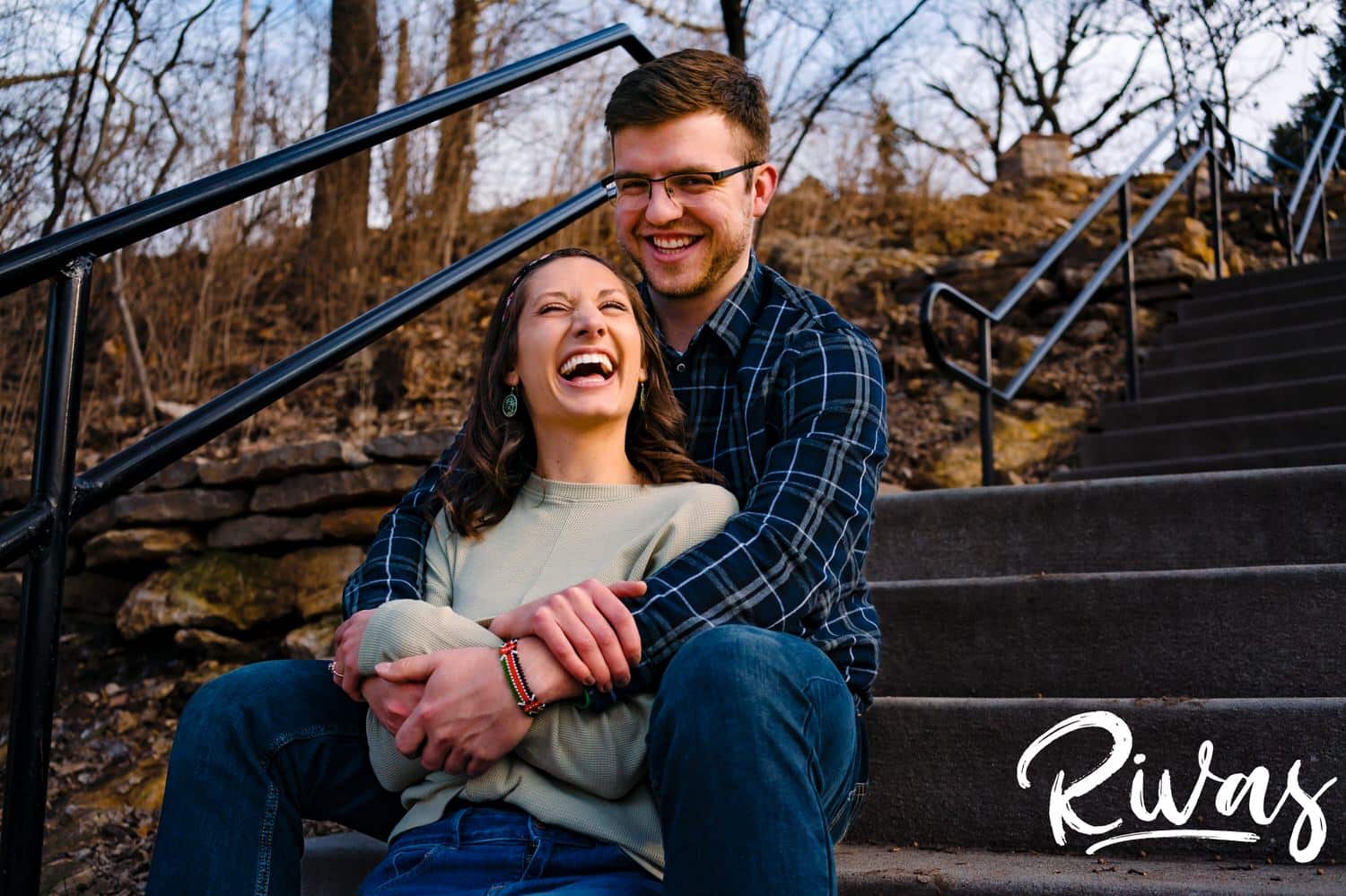 A relaxed, candid picture of an engaged couple sitting on stairs, sharing an embrace and laughing together during their winter engagement session in Kansas City. 