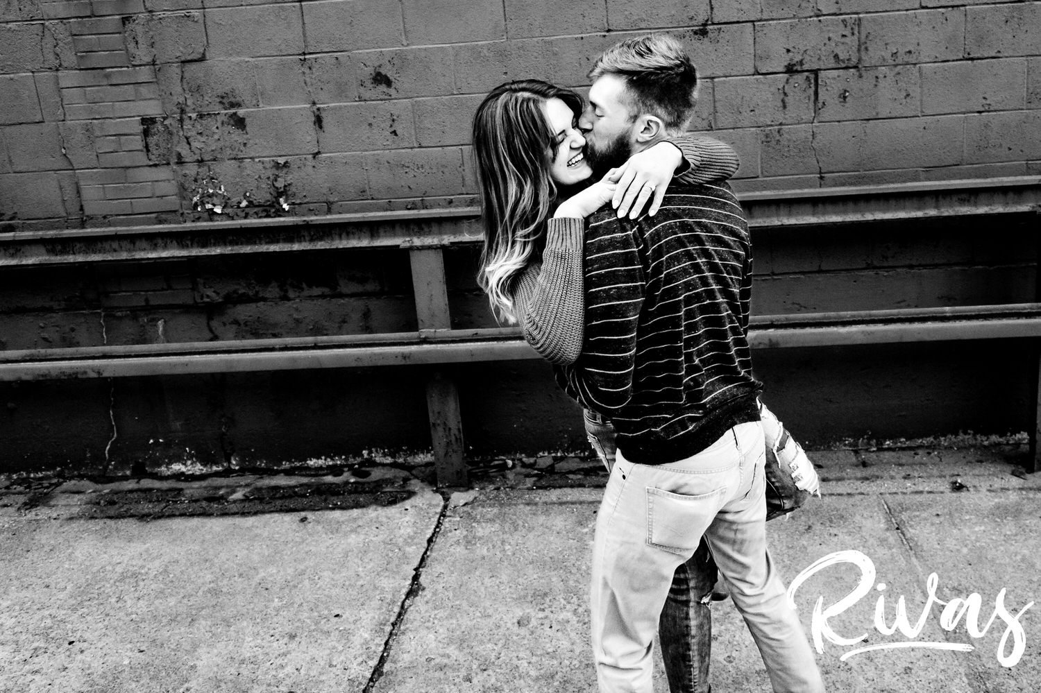 A candid, black and white picture of an engaged couple laughing and sharing an embrace together in Kansas City's West Bottoms neighborhood during their winter engagement session.