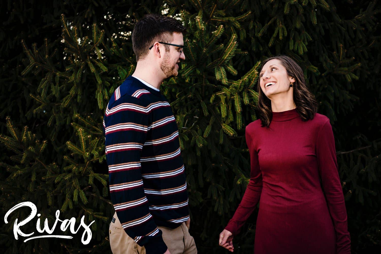 A colorful, candid picture of an engaged couple laughing together in front of a wall of green trees during their winter engagement session at The Nelson Atkins Museum of Art.
