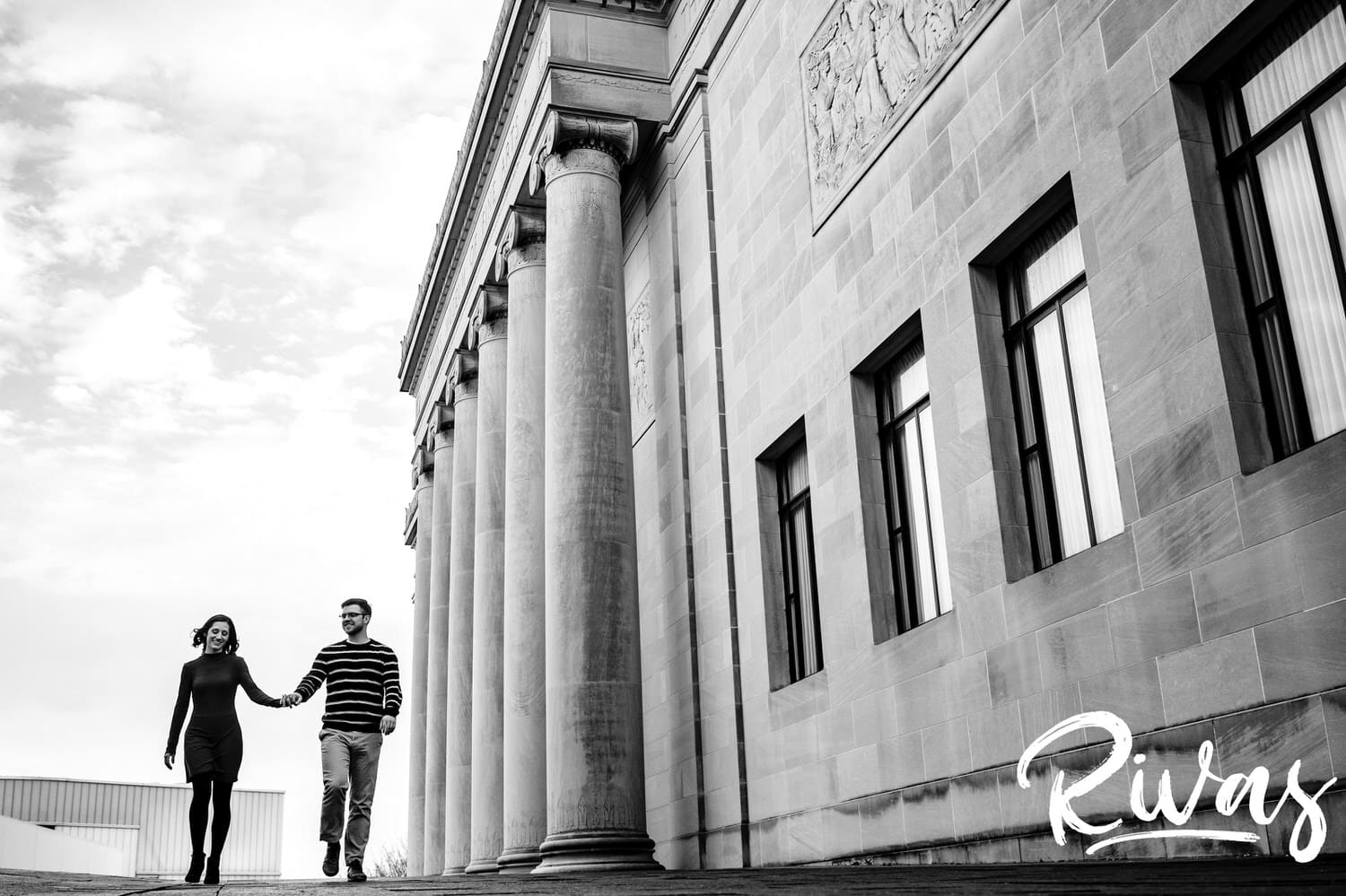 A wide, candid picture of a couple walking hand-in-hand in front of the iconic columns at The Nelson Atkins Museum of Art during their winter engagement session. 