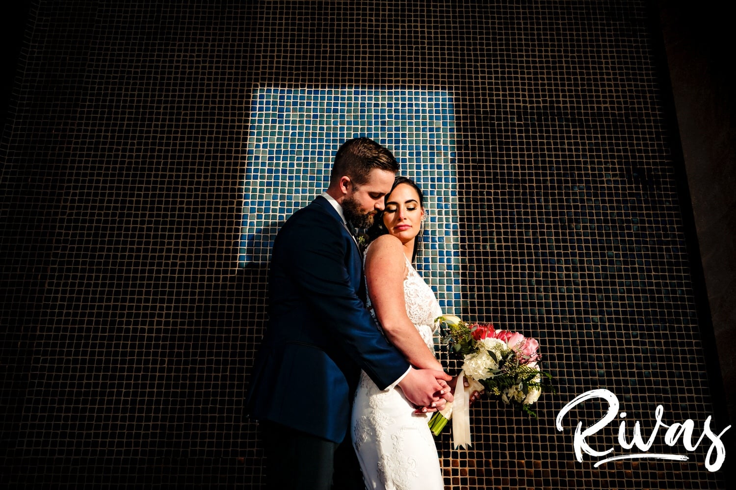 A bold, colorful picture of a modern bride and groom sharing an embrace up against a blue tiled wall bathed in a bright spot of sunlight during their winter wedding day in Kansas City.