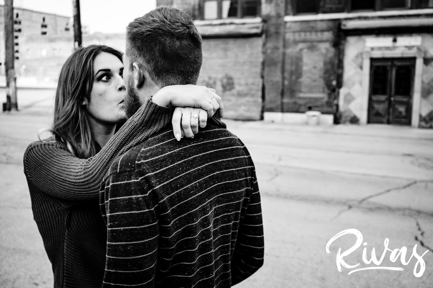 A silly, black and white picture of a woman sticking her tongue out and crossing her eyes at her fiance as she wraps her arms around his neck during their engagement session in the West Bottoms. 