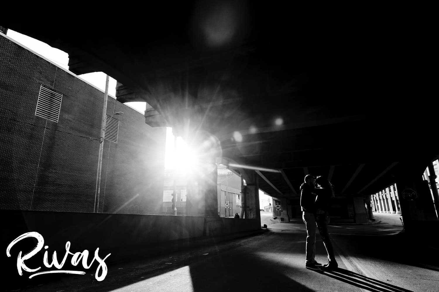A candid black and white, wide picture of an engaged couple dancing together under the 12th Street Bridge in Kansas City's West Bottoms as sunlight bathes them in bright light during their engagement session. 