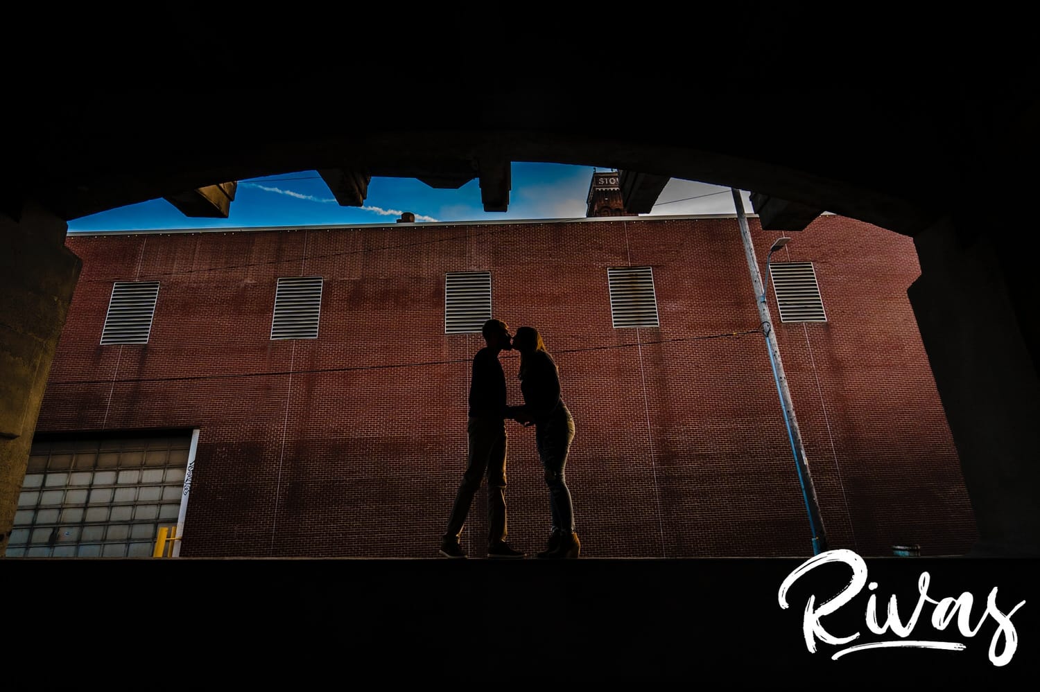 A colorful silhouetted picture of an engaged couple standing on a ledge in front of a brick wall, leaning in to share a kiss during their engagement session in the West Bottoms in Kansas City. 