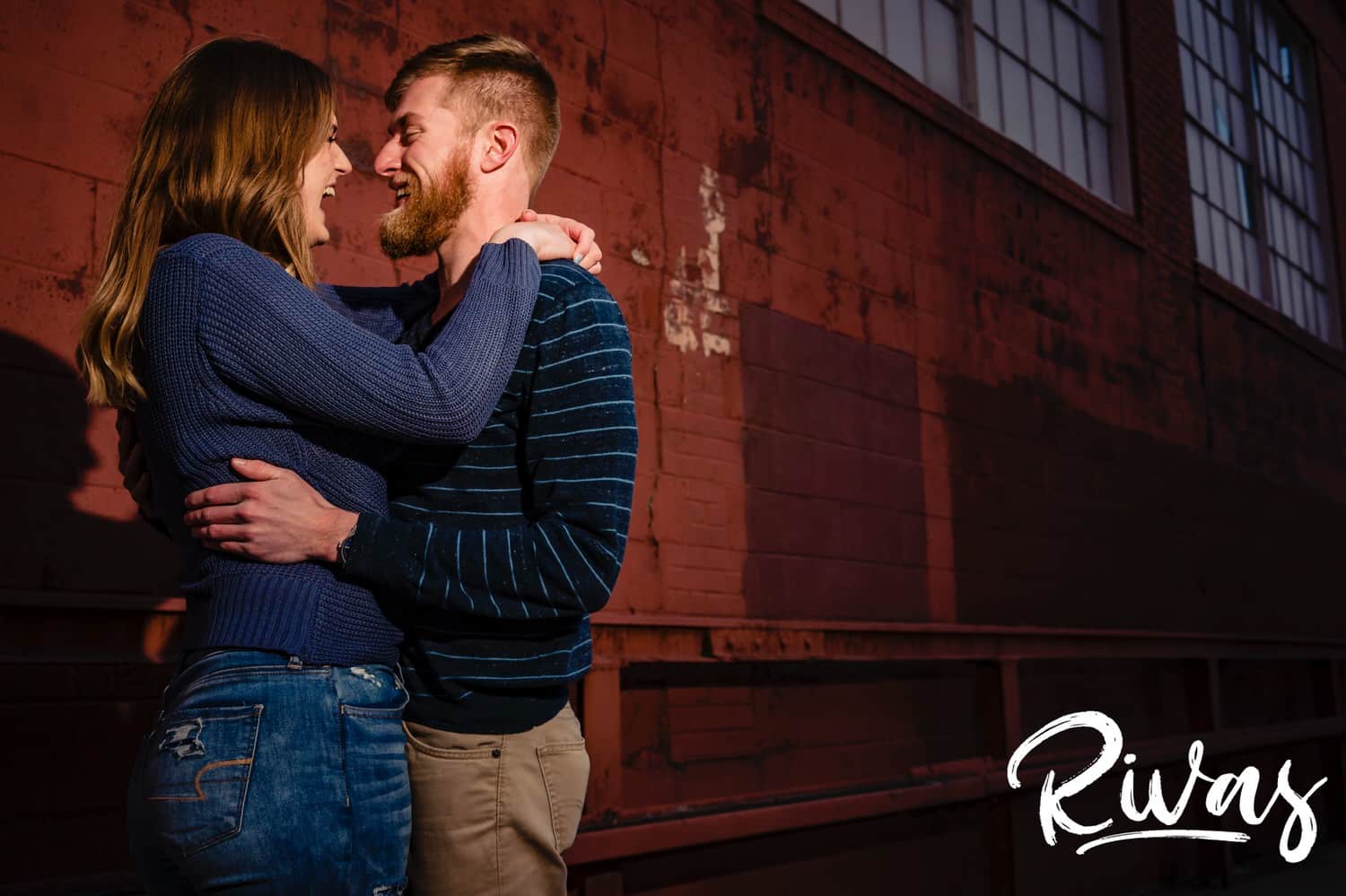 A colorful, intimate picture of an engaged couple standing face to face, laughing together as they stand in front of a red wall during their engagement session in Kansas City's West Bottoms. 
