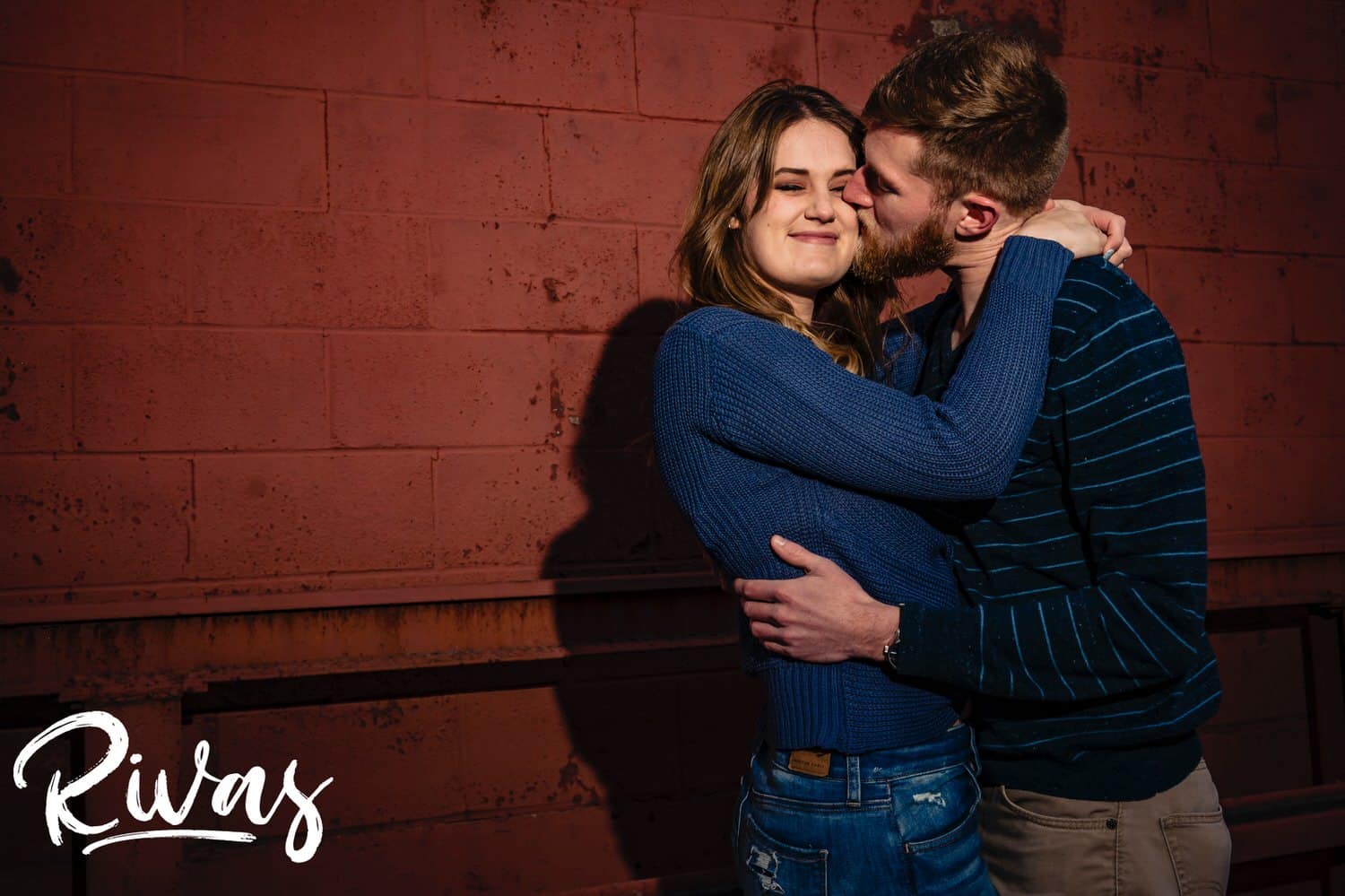 A colorful, intimate picture of an engaged couple standing face to face, laughing together as they stand in front of a red wall during their engagement session in Kansas City's West Bottoms. 