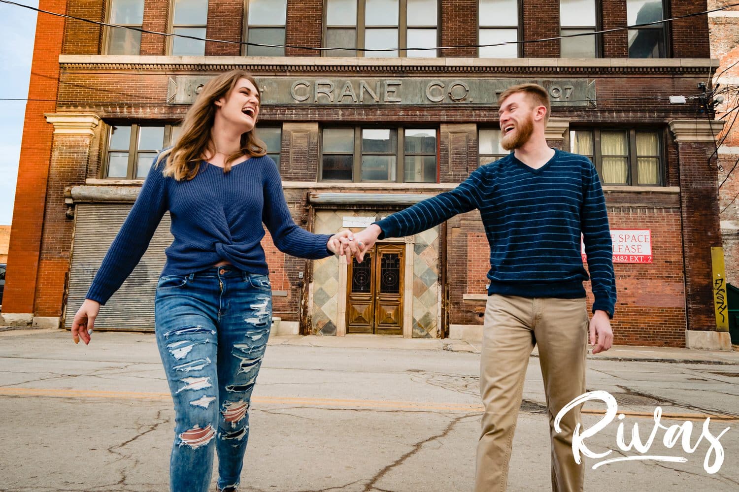 A candid picture of an engaged couple holding hands and laughing together as they walk down the middle of the street during their West Bottoms Engagement Session in Kansas City. 