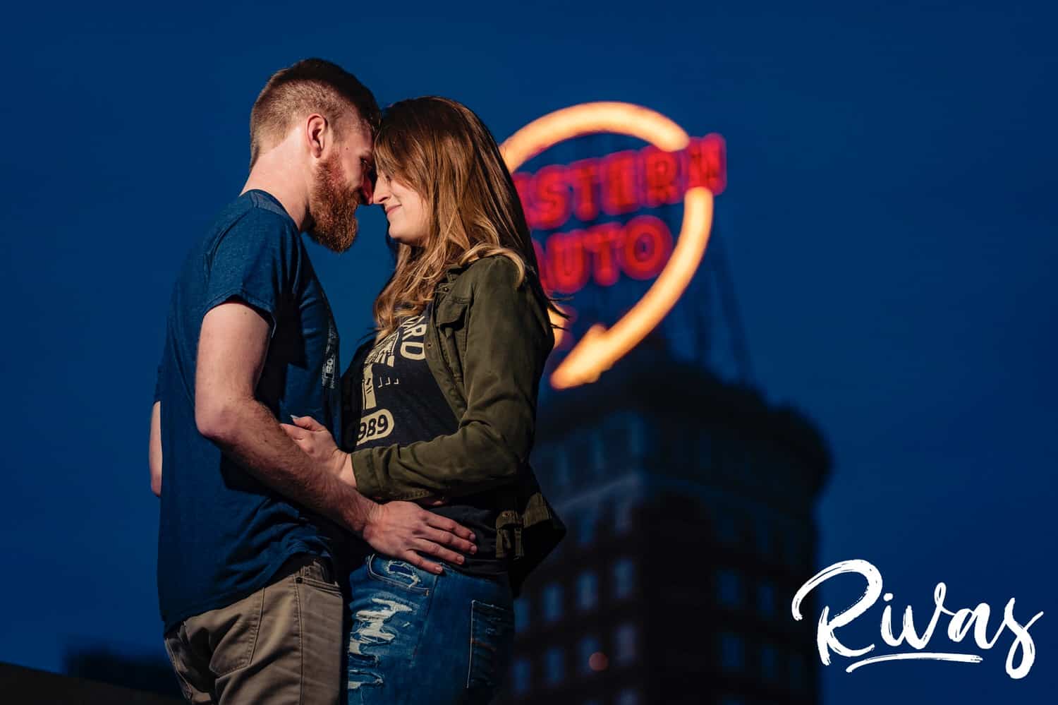 A candid picture of an engaged couple walking hand in hand in front of the lit-up Western Auto sign during their downtown Kansas City engagement session. 