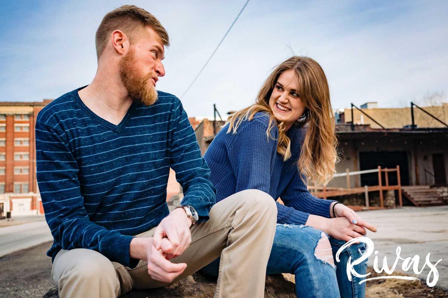 A casual, colorful picture of an engaged couple sitting on a pile of rocks laughing together in the middle of Kansas City's West Bottoms during their engagement session. 