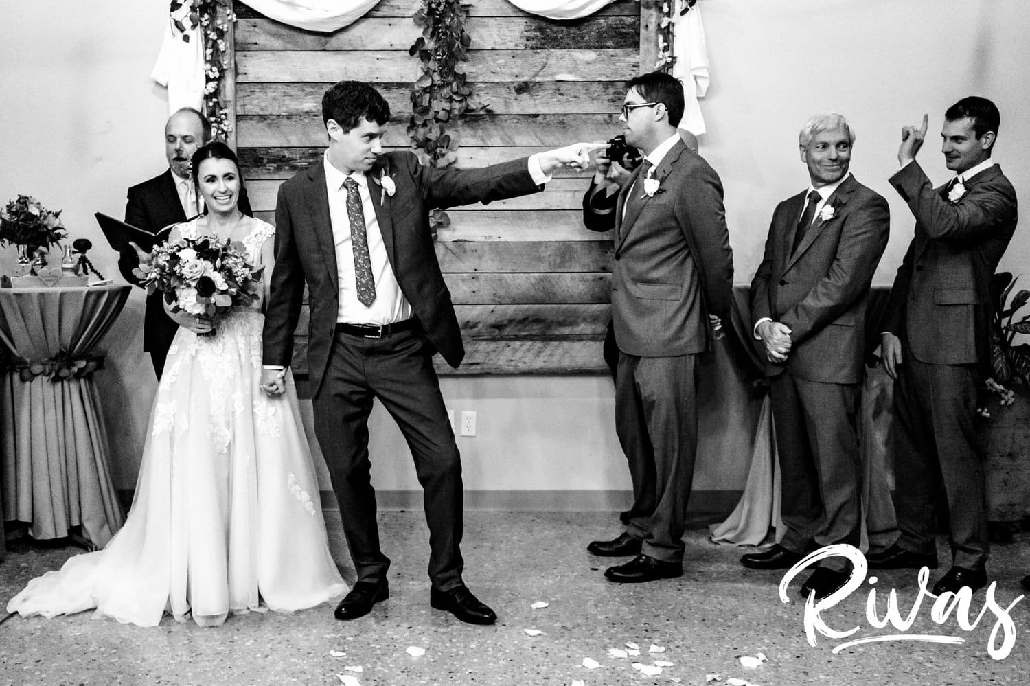 A candid, black and white picture of a groom laughing and pointing at his mom in the audience during the middle of his rainy day wedding ceremony at The Bowery in Kansas City. 