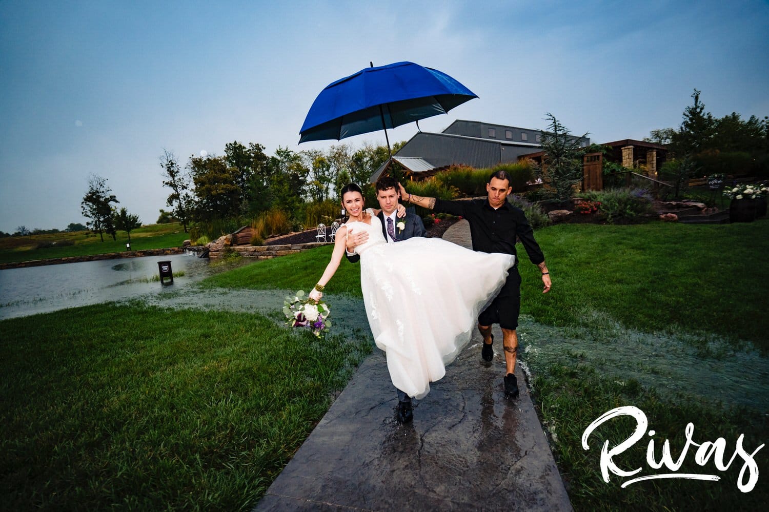 A colorful picture of a groom carrying his bride through a stream of water as they enter their reception on a rainy wedding day at The Bowery in September. 