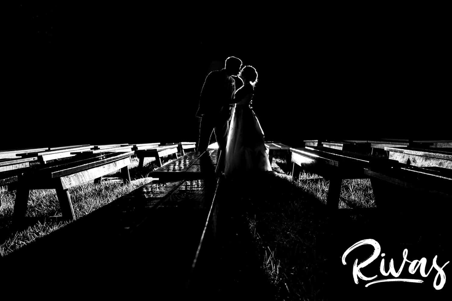A dramatic portrait of a silhouetted bride and groom standing between wooden benches covered in shiny water at the end of their wedding day at The Bowery in Kansas City. 
