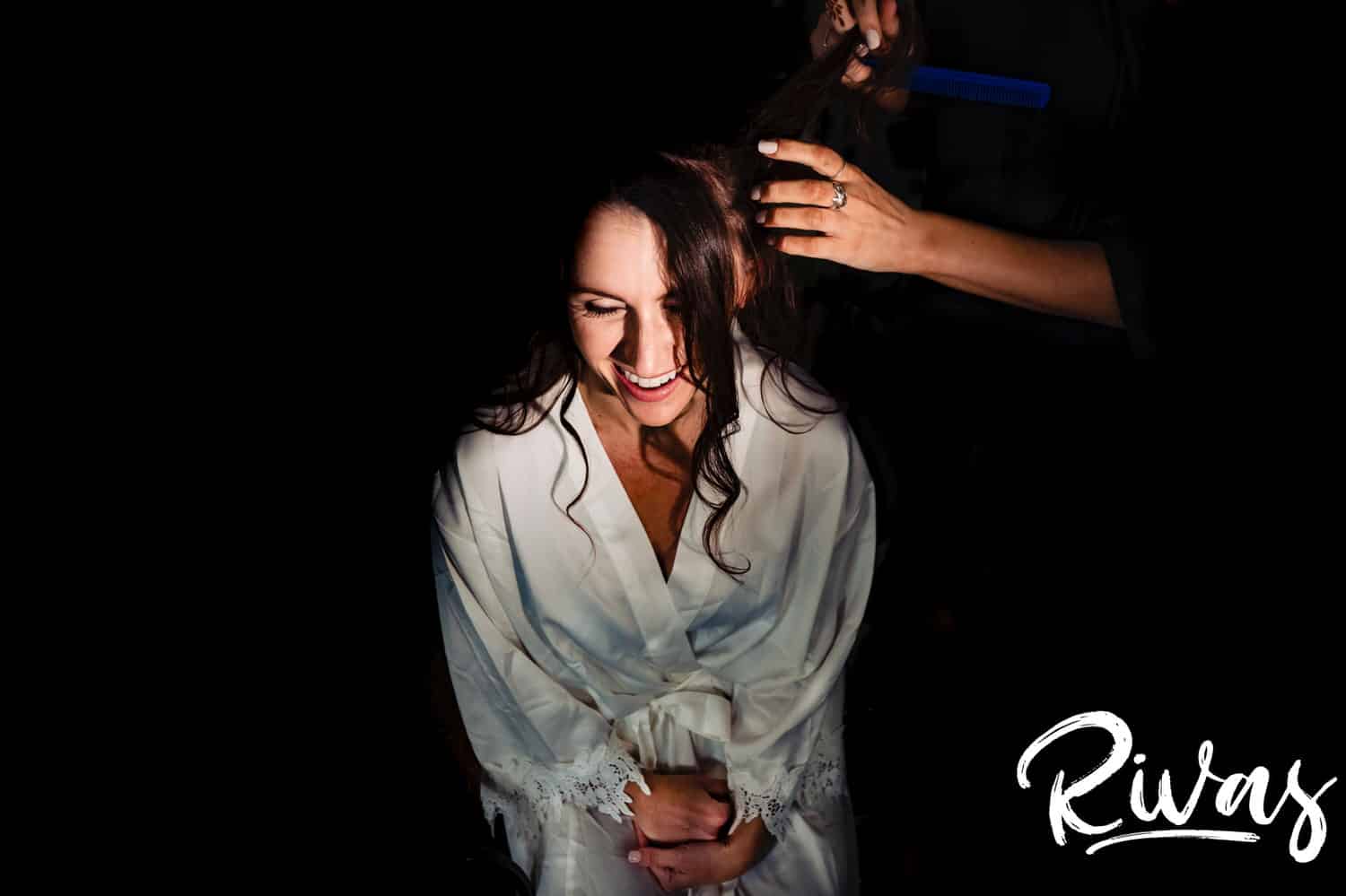 A dark, candid picture of a bride clasping her hands together as a hairdresser curls her hair on the morning of her wedding at The Bowery in Kansas City. 