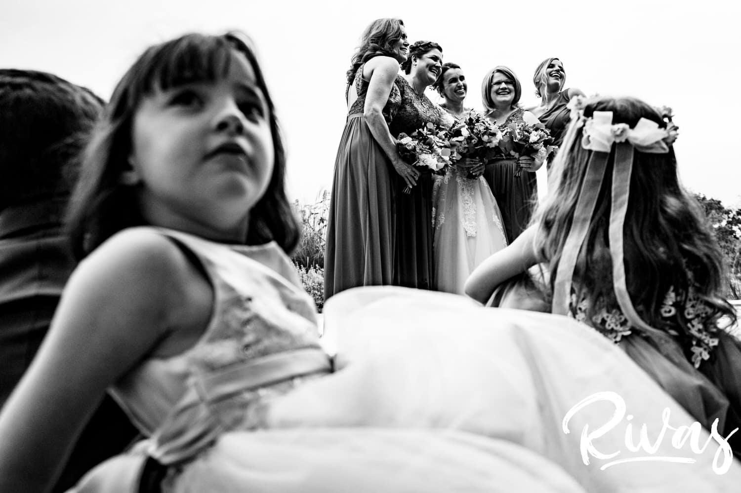 A candid black and white picture taken from behind the shoulder of a flower girl of a bride and her bridesmaids having their photo taken on the afternoon of a rainy wedding day at The Bowery. 