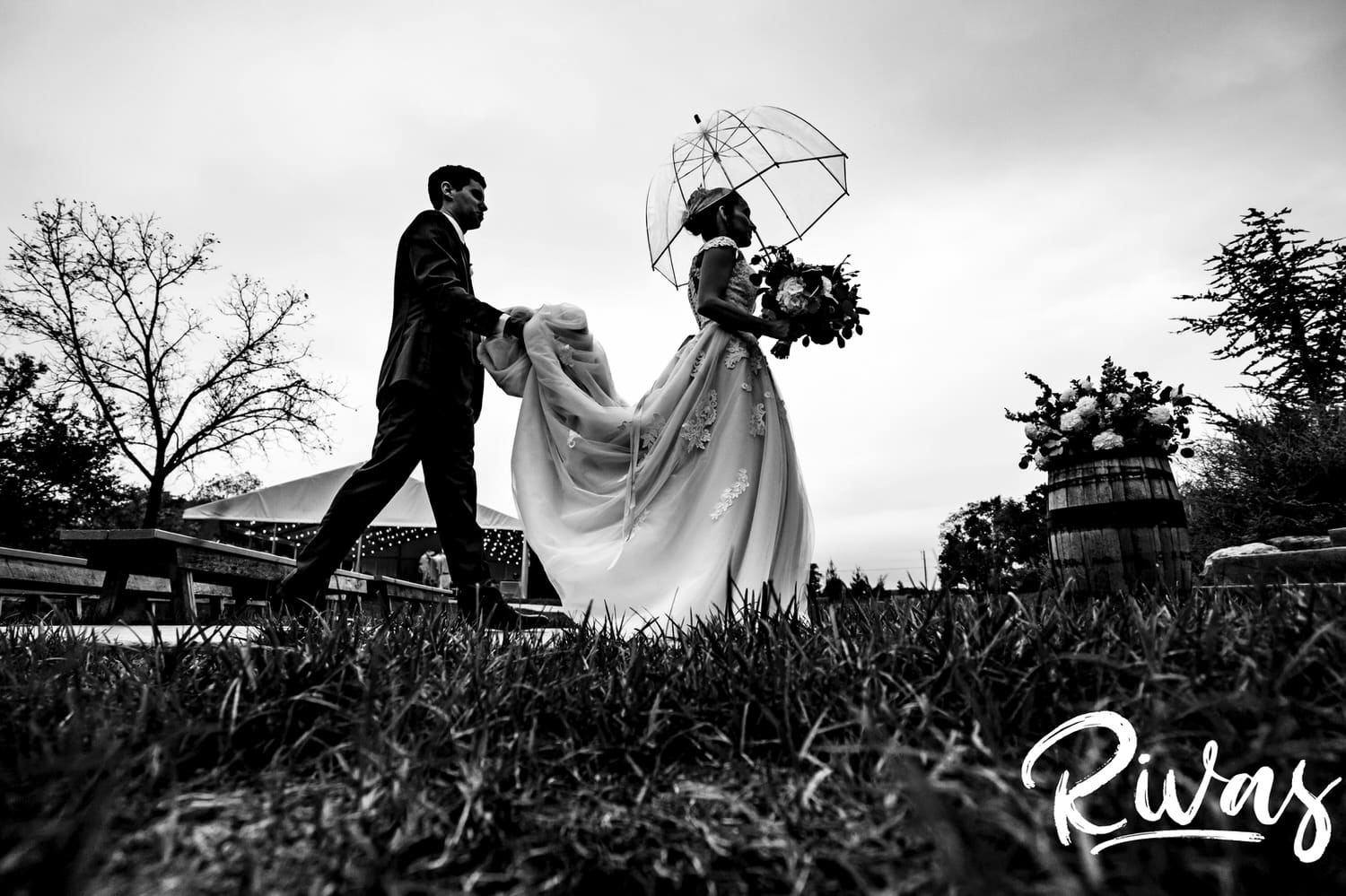 A candid black and white picture of a bride holding her bouquet and a clear umbrella as her groom follows her, holding up her train for her on their rainy wedding day in Kansas City at The Bowery. 