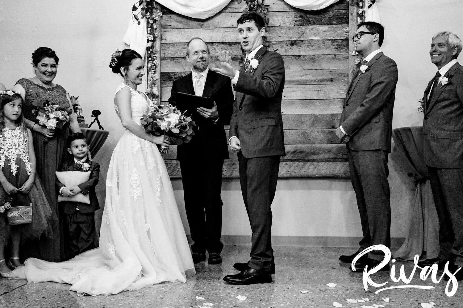 A candid, black and white picture of a groom laughing and pointing at his mom in the audience during the middle of his rainy day wedding ceremony at The Bowery in Kansas City. 