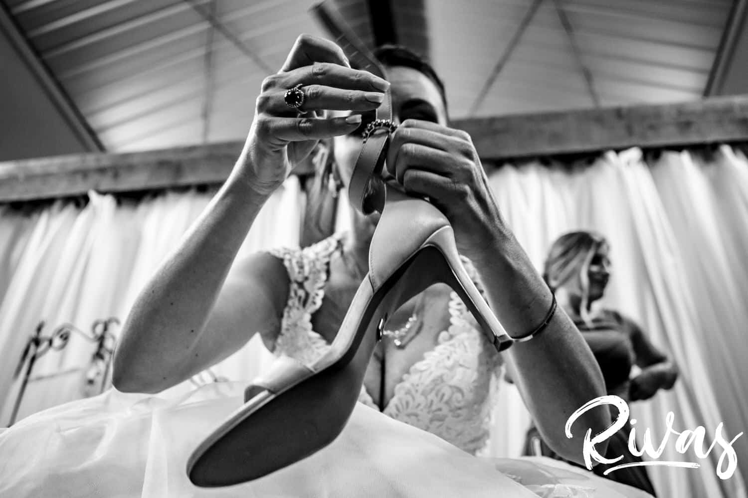 A candid black and white picture of a bride holding up her shoe as she tries to put it on. 