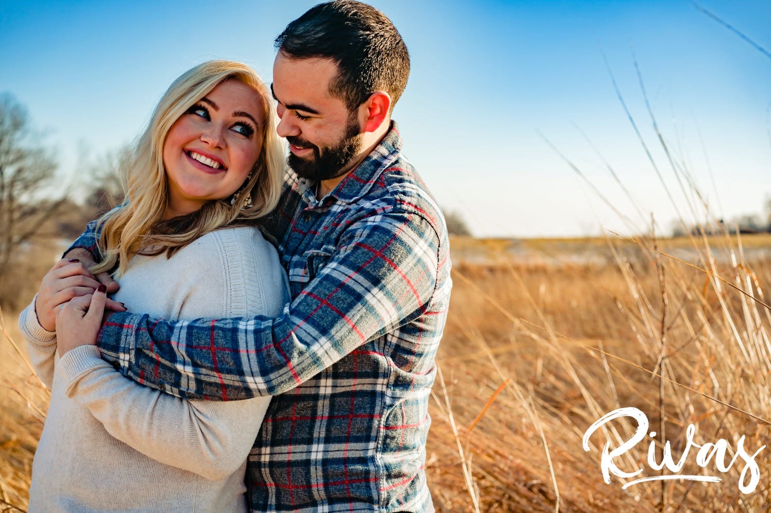 A bright, candid picture of an engaged couple sharing an embrace and laughing together as they stand in a field during their bright winter engagement session in Kansas City. 