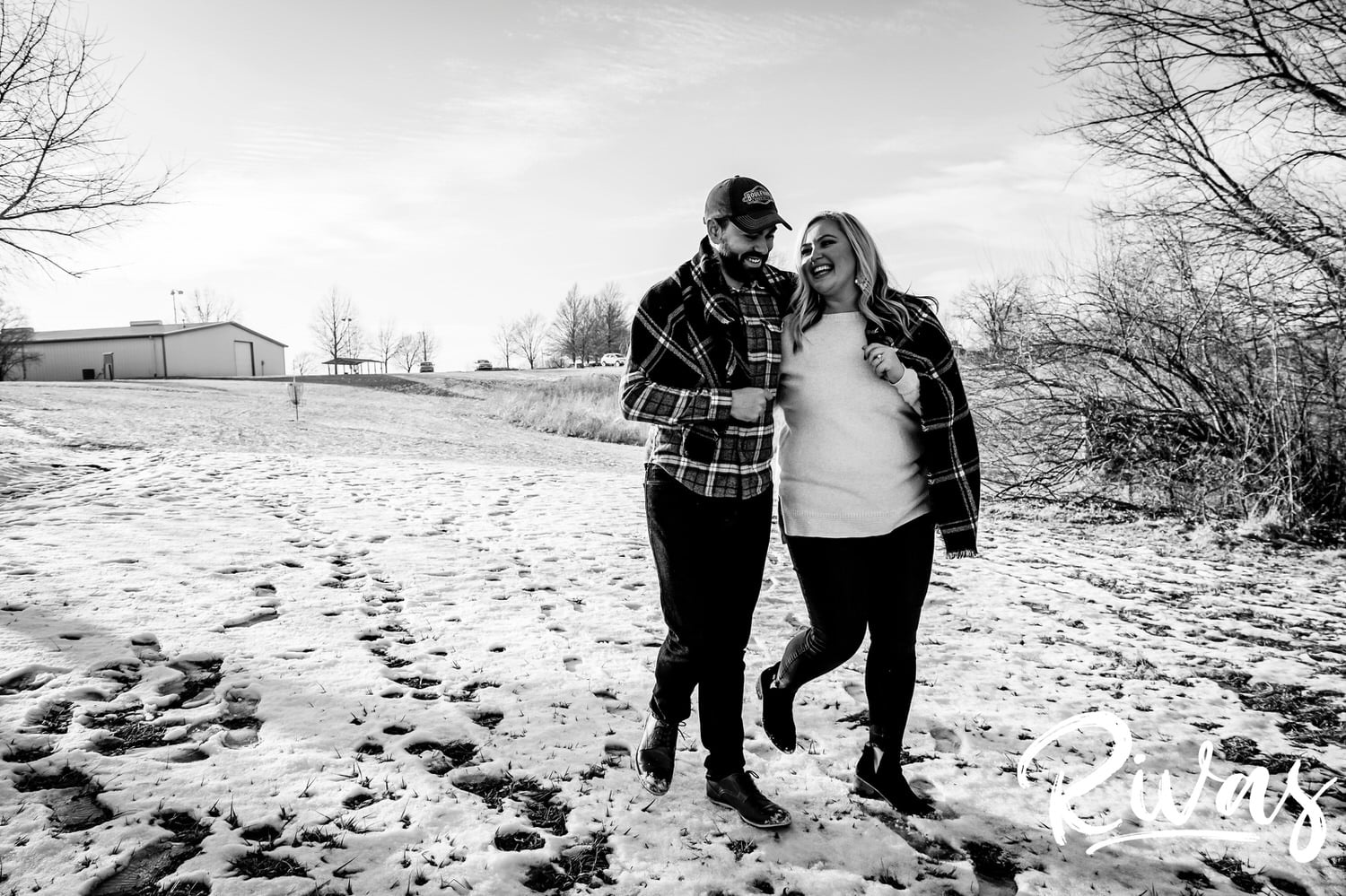 A candid black and white picture of an engaged couple sharing an embrace as they walk through a snowy field during their winter engagement session in Kansas City. 