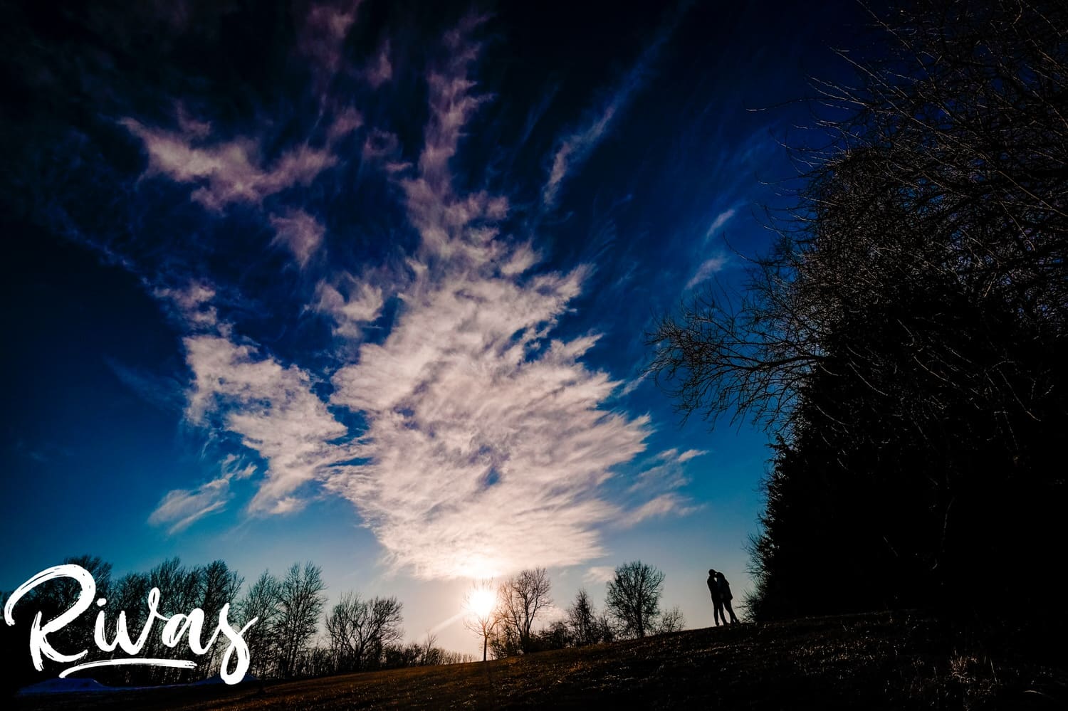 A vibrant, wide, silhouetted picture of an engaged couple standing on a hilltop against a bright blue sky during their winter engagement session in Kansas City. 