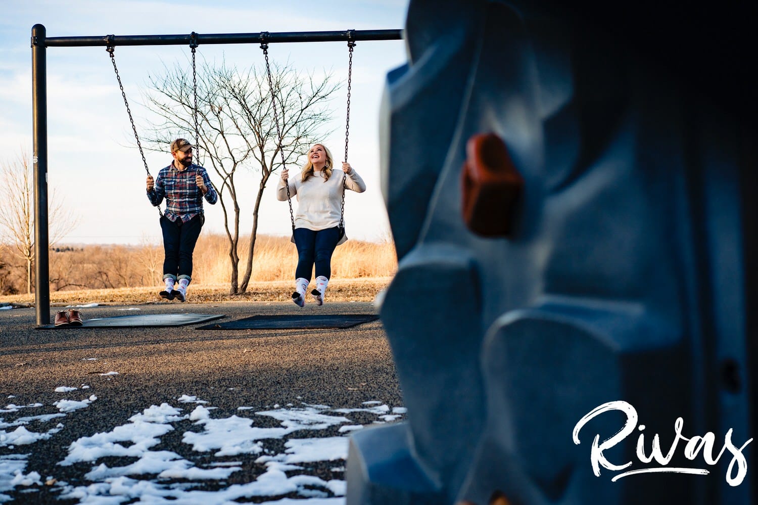 A wide, candid picture of an engaged couple swing on a swing set in their socks during their winter engagement session. 