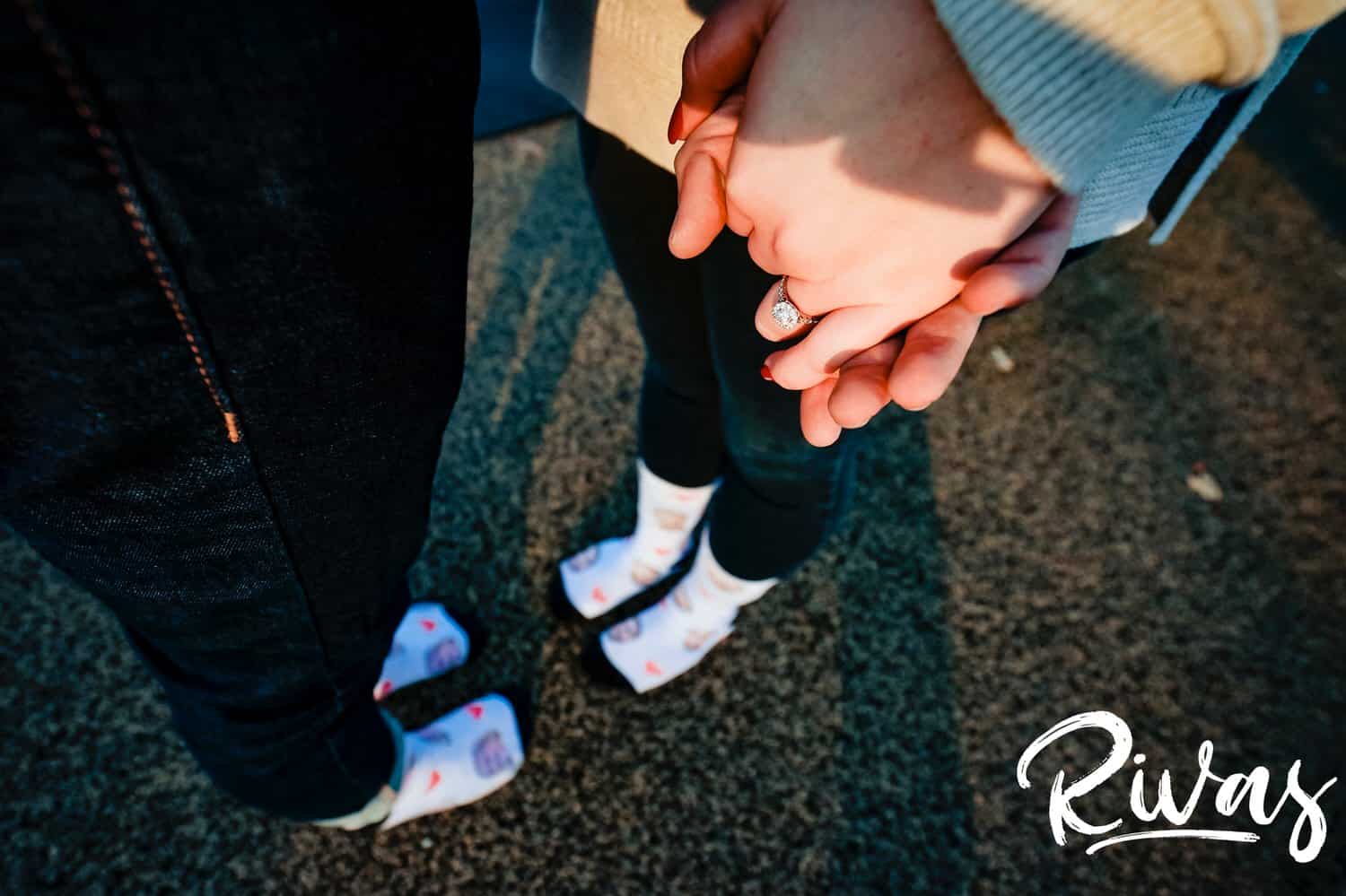 A tight, detailed picture of an engaged couple holding hands, with the picture focused on an engagement ring and their socks visible in the background. 
