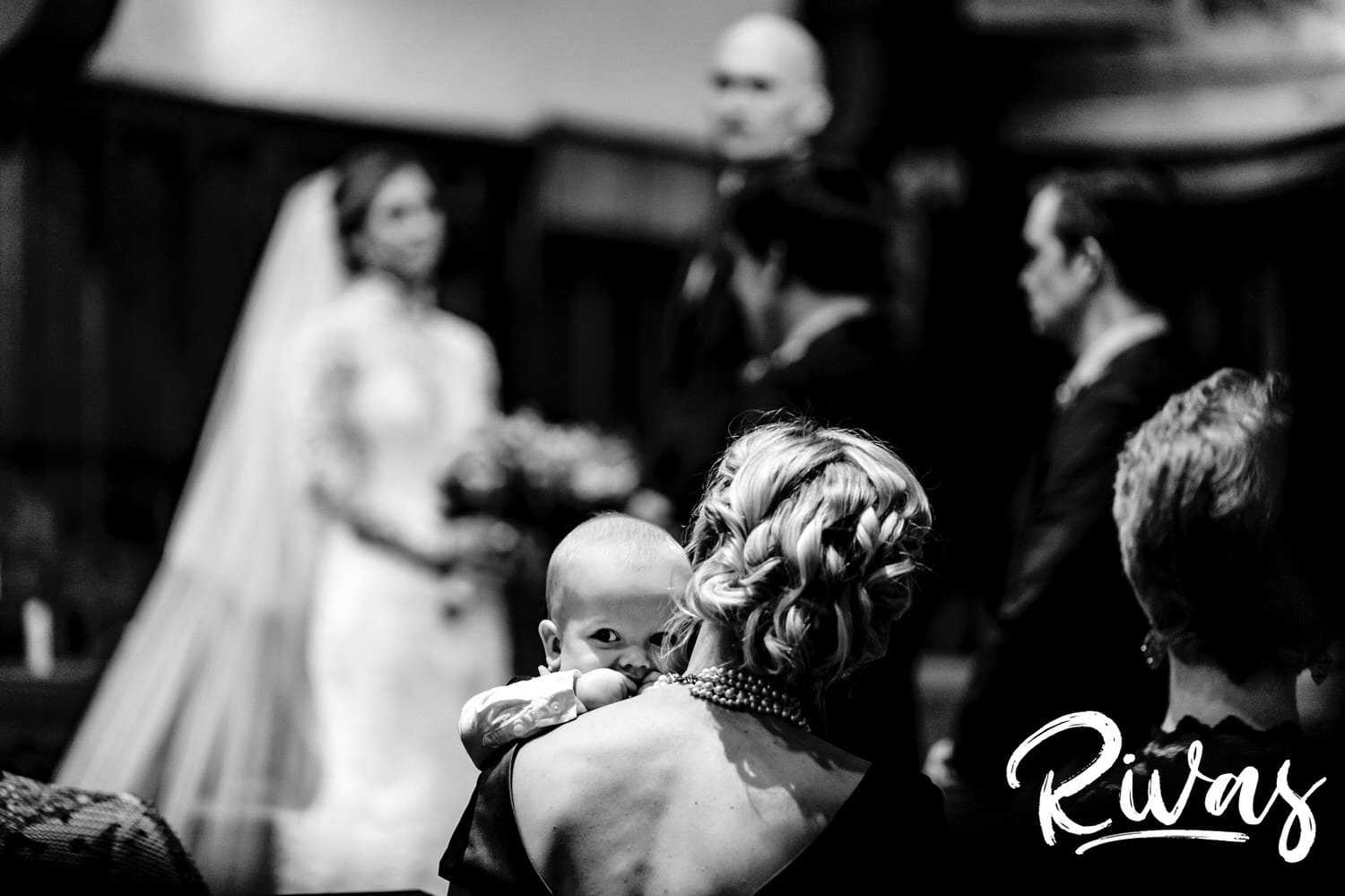 A candid black and white picture taken from a distance of a baby ring bearer chewing on his mom's pearls during a wedding ceremony with the bride and groom visible in the background. 
