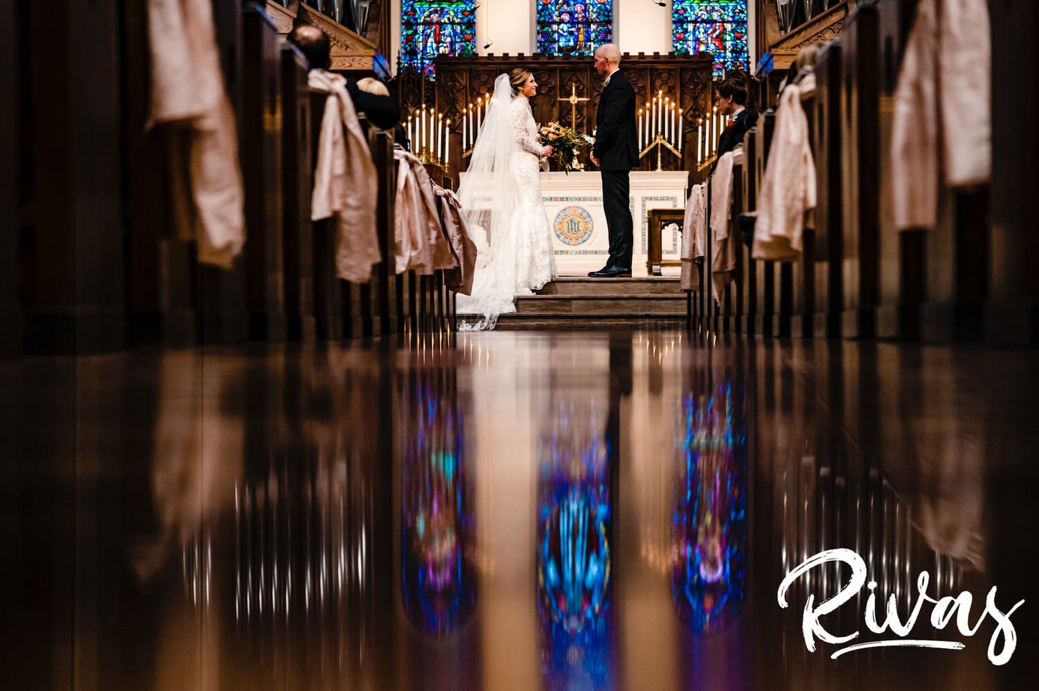 A colorful, wide picture taken from the ground looking up of a bride and groom holding hands at the front of the church with the church's stained glass windows reflected in the shiny floor. 