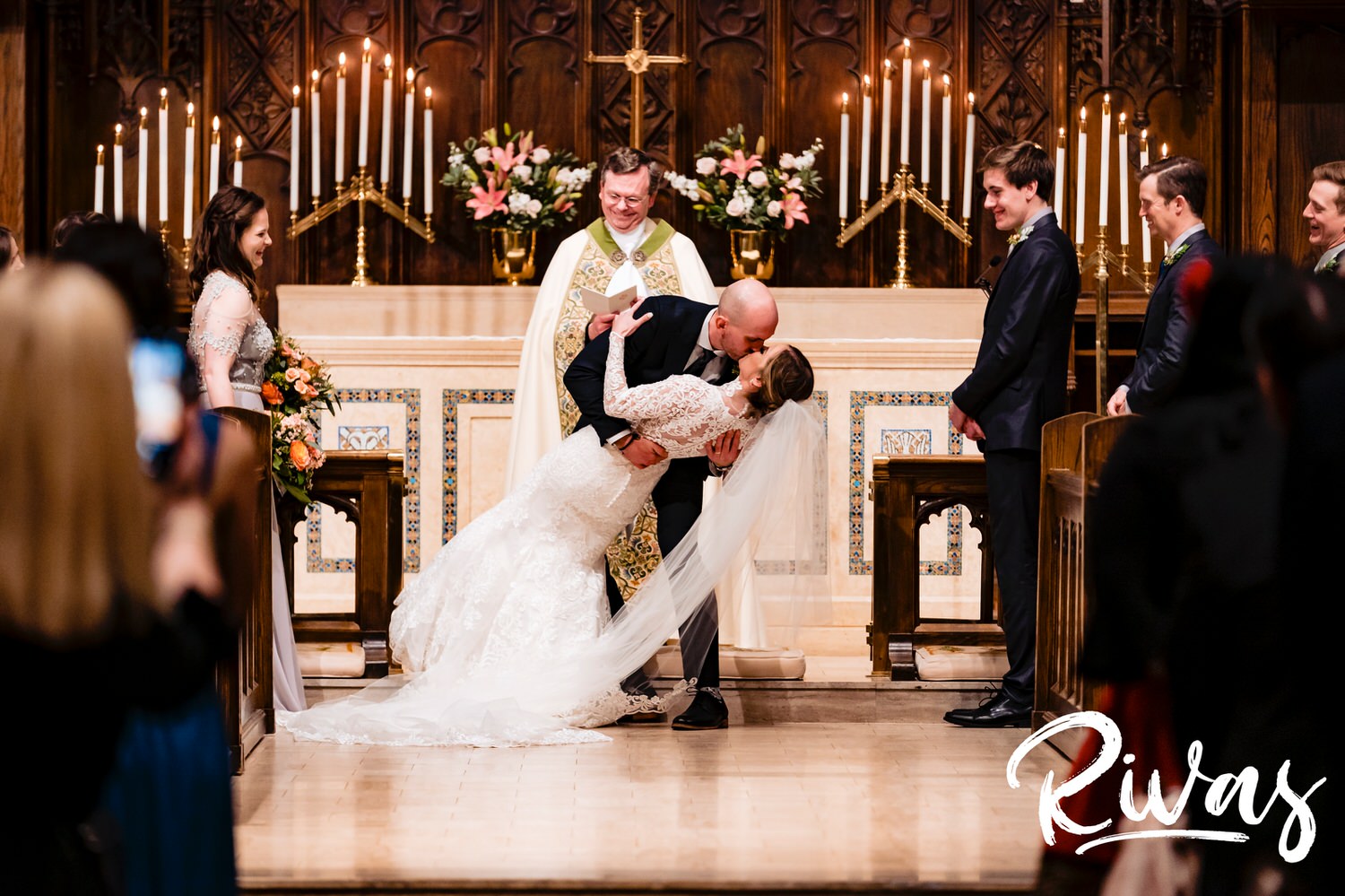 A colorful, wide image taken from the back of the church as a groom dips his bride back during their first kiss at the end of their wedding ceremony. 