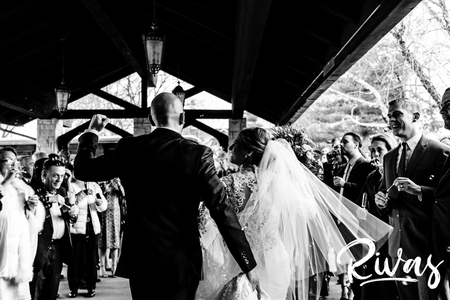 A candid black and white picture taken from behind of a bride looking lovingly up at her groom, celebrating his hands in celebration as they exit the church under a flurry of bubbles on their wedding day. 