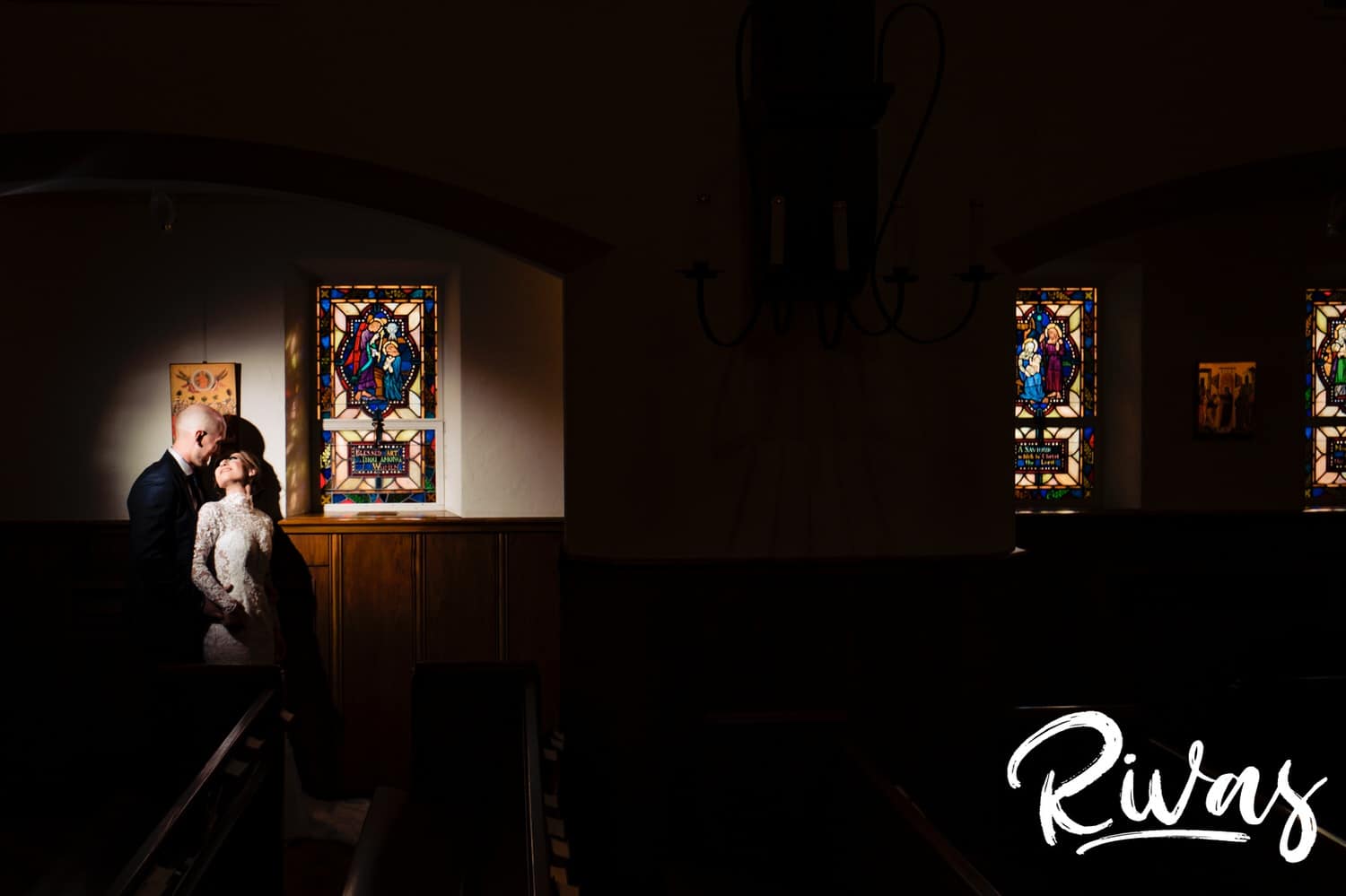 A dark and dramatic portrait of a bride and groom leaning up against a wood paneled wall just in front of a stained glass window at St. Andrew's Episcopal Church in Kansas City on a sunny winter wedding day. 