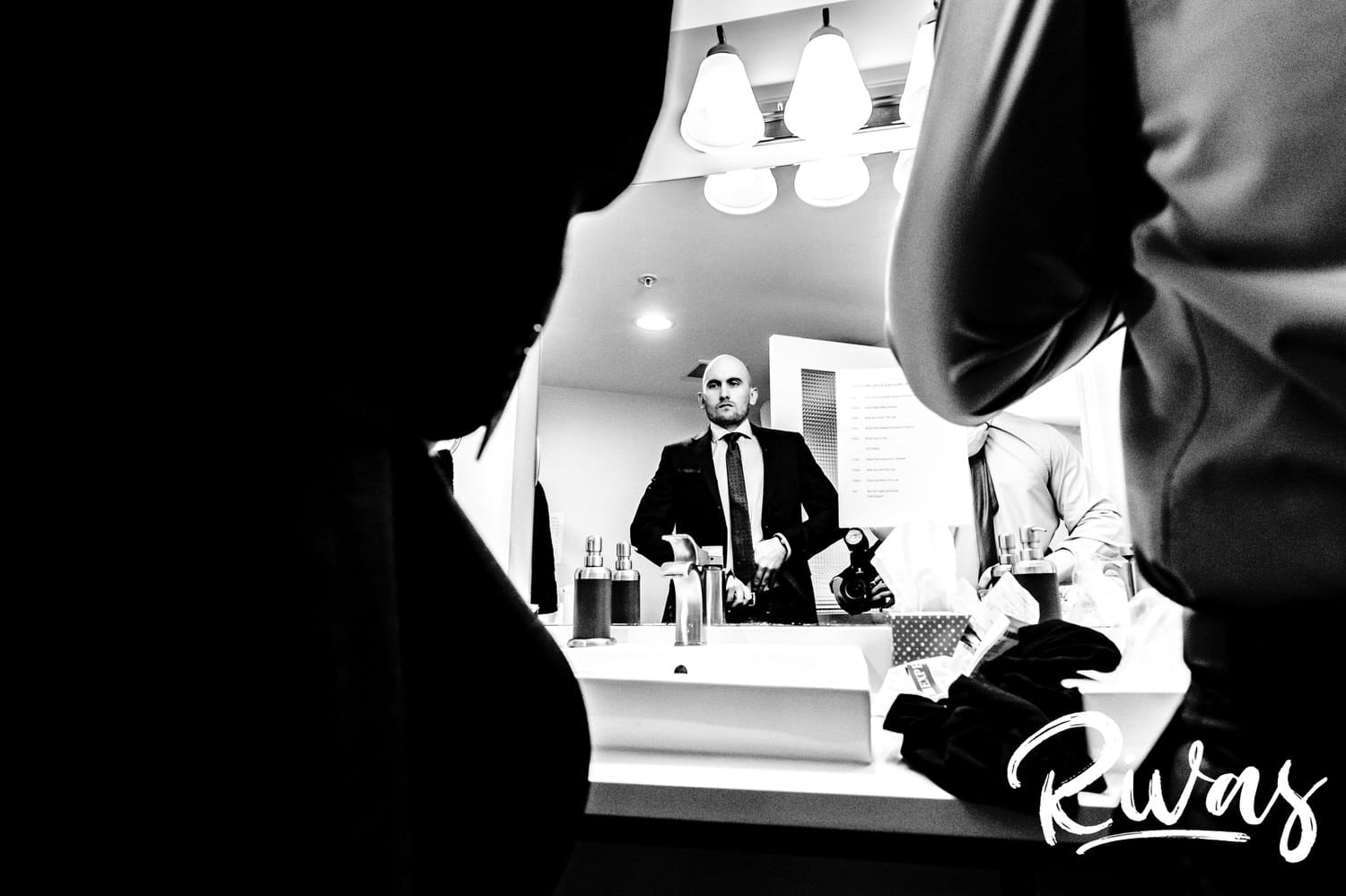 A candid black and white picture taken from between two men of a man in a dark suit checking his reflection before he sees his bride for the first time on their wedding day. 