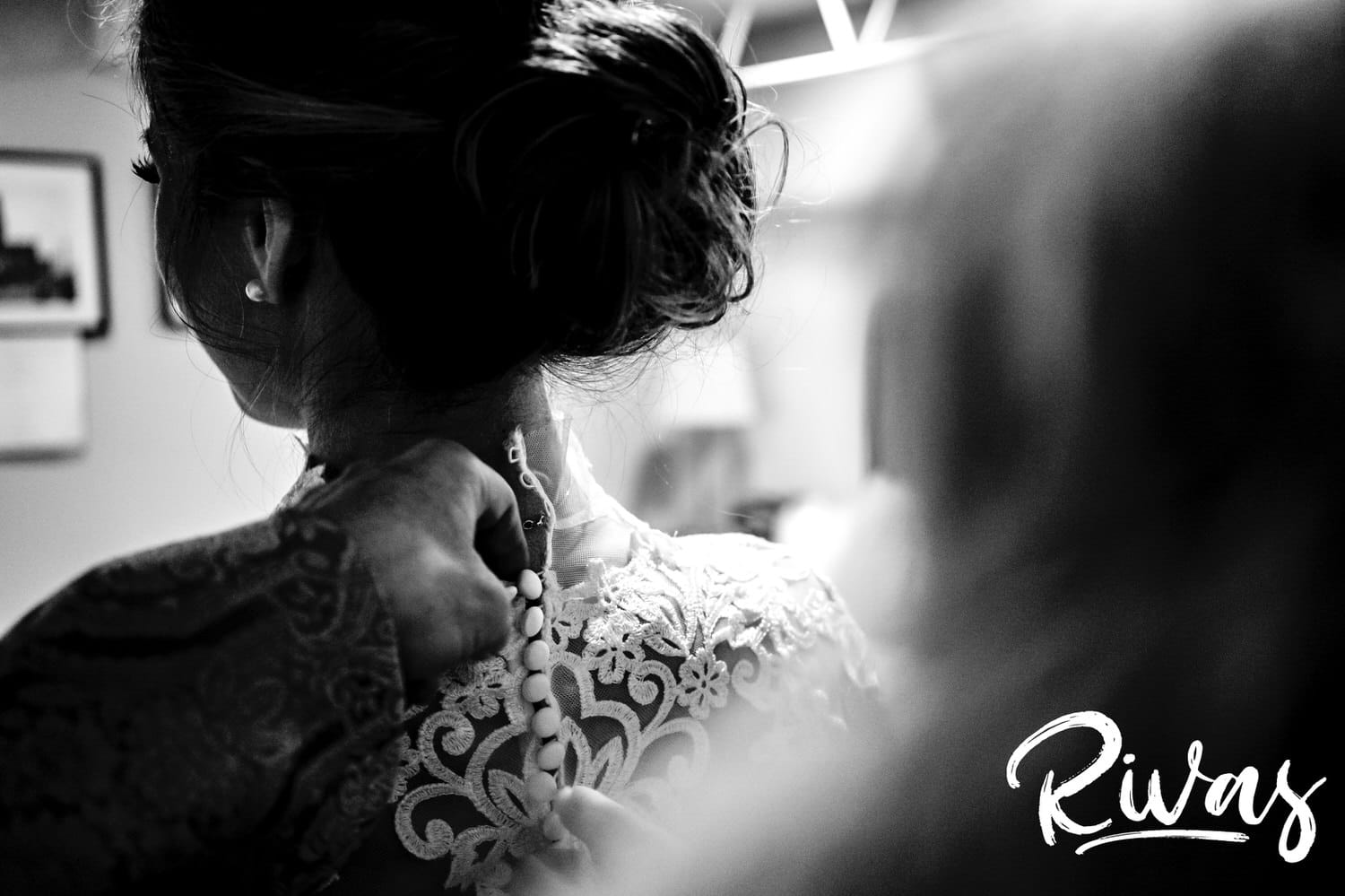 A candid, tight, black and white picture taken over the shoulder of a bride's mom buttoning up the buttons on her daughter's wedding gown on the morning of her sunny winter wedding. 