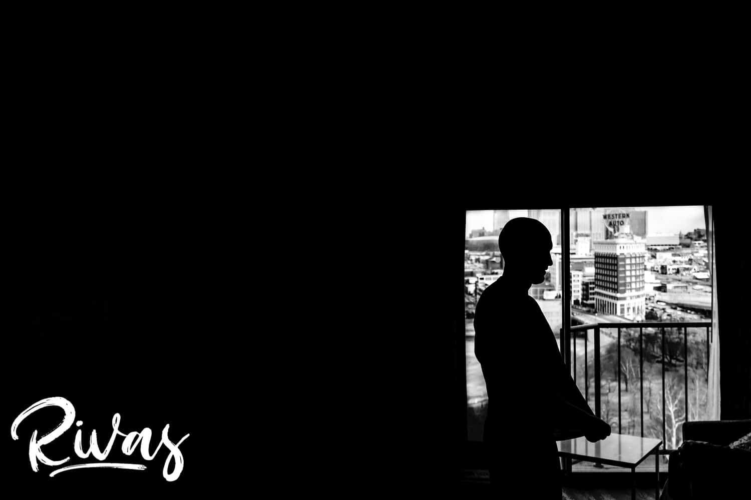 A dramatic black and white candid picture of a man standing in the bright window of a hotel room, looking out over the Kansas City skyline on the day of his winter wedding. 