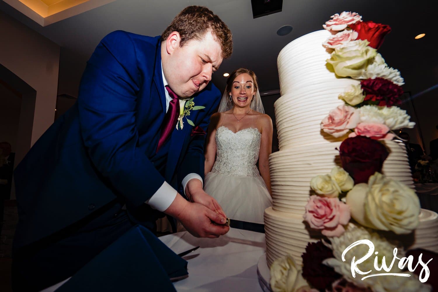 A candid picture of a groom cutting through two layers of wedding cake with a very serious look on his face as his bride surprisingly looks on in the background during a wedding reception at Piazza Messina. 