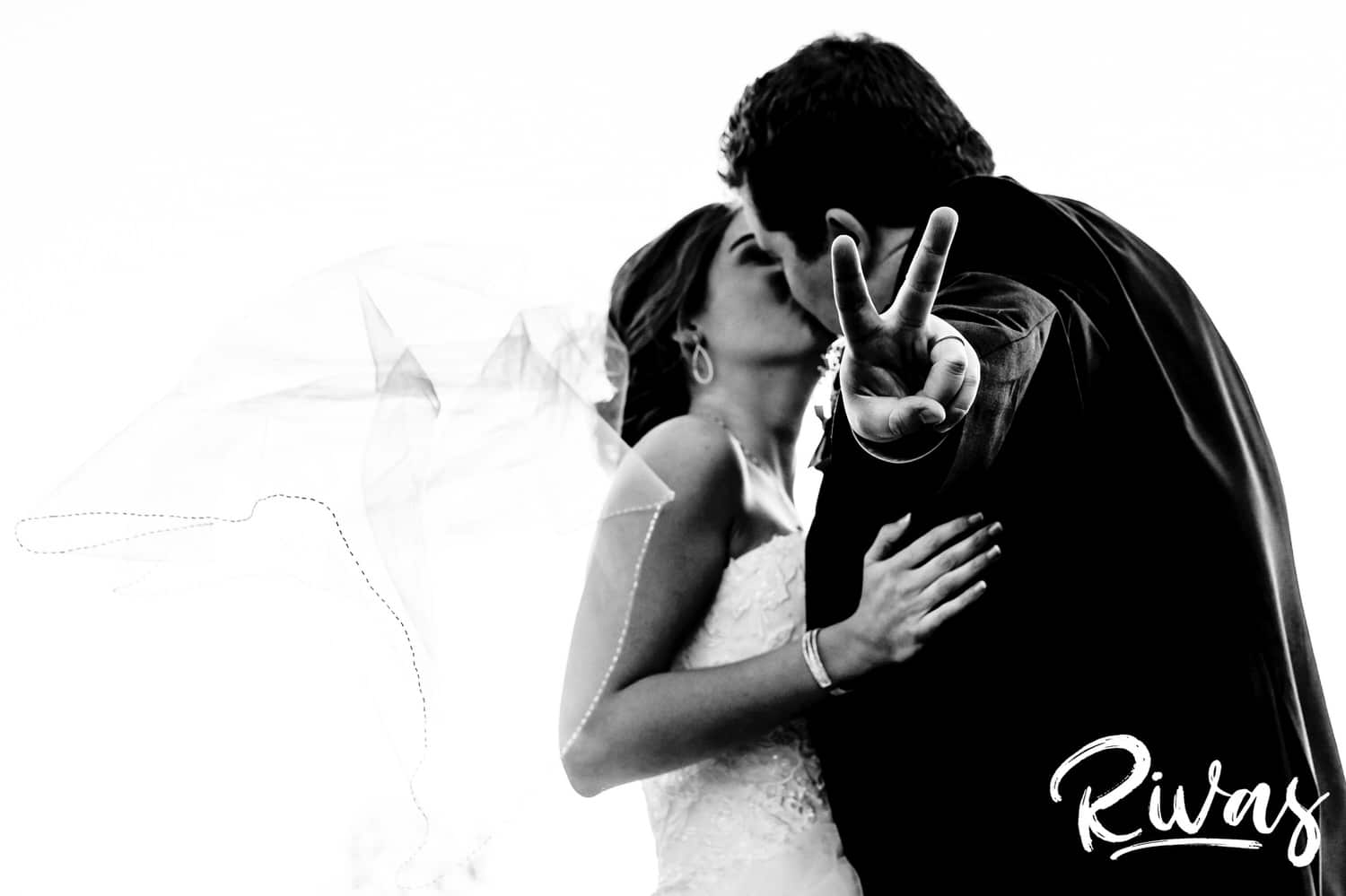A black and white picture of a bride and groom leaning in to share a kiss as the groom throws up a peace sign at the camera. 
