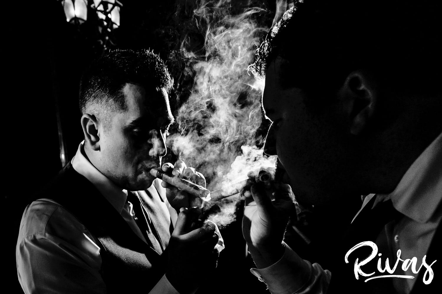 A dramatic black and white picture of a groomsman trying to light another groomsman's cigar during a fall wedding reception. 